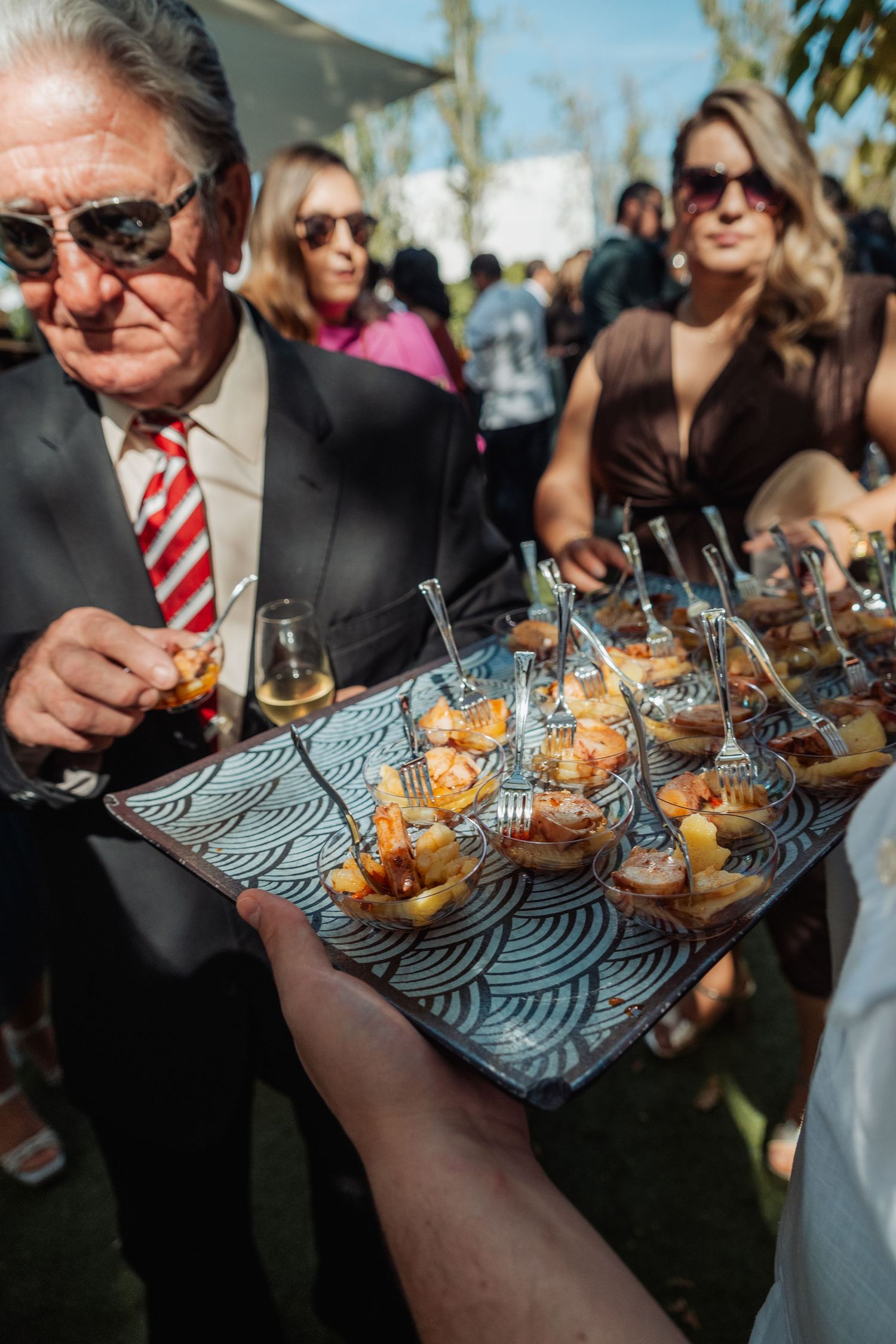 Wedding guest in suit examines appetizer tray with shrimp canapés at outdoor reception.