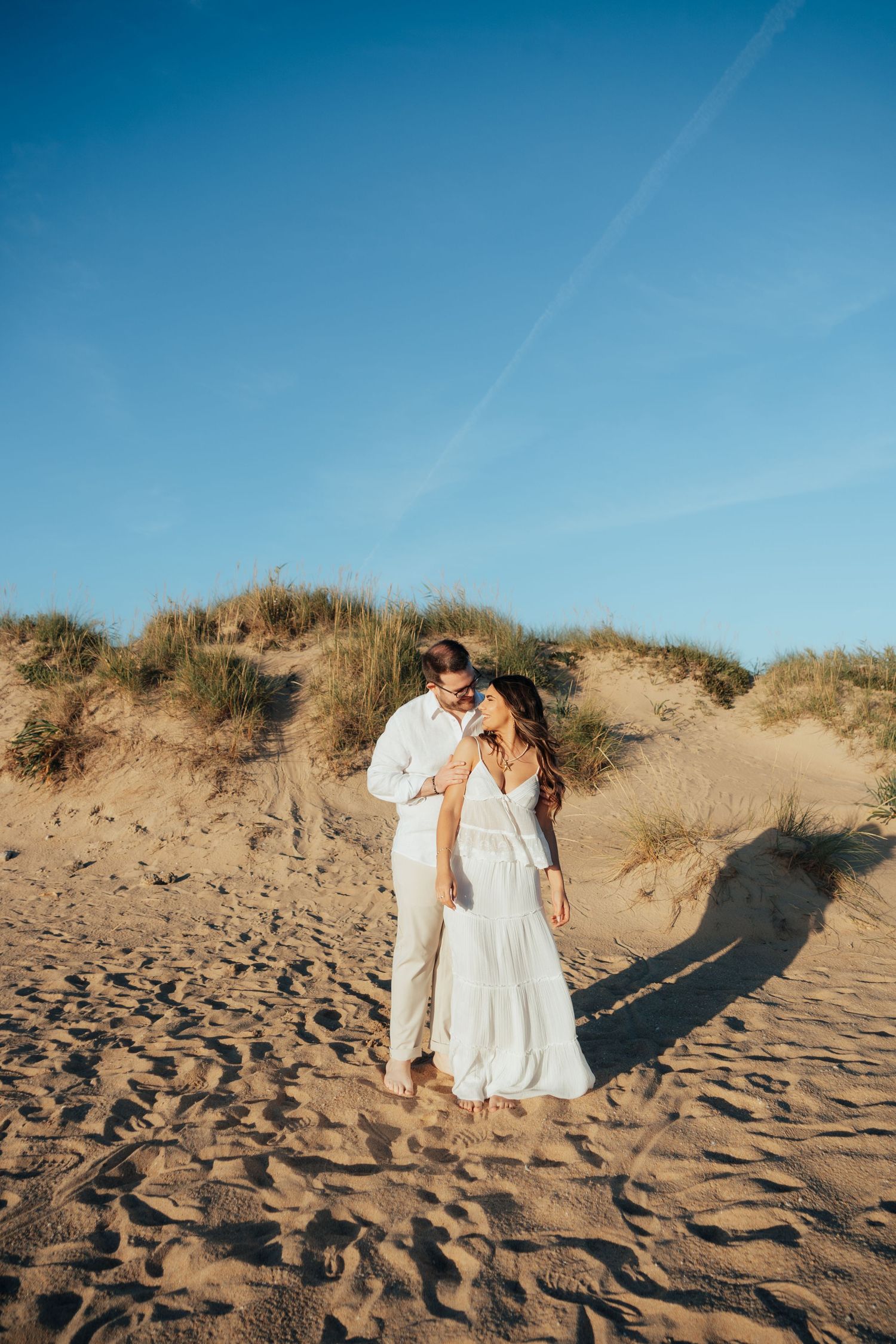 Couple in white outfits embracing on sandy beach dunes under a clear blue sky.