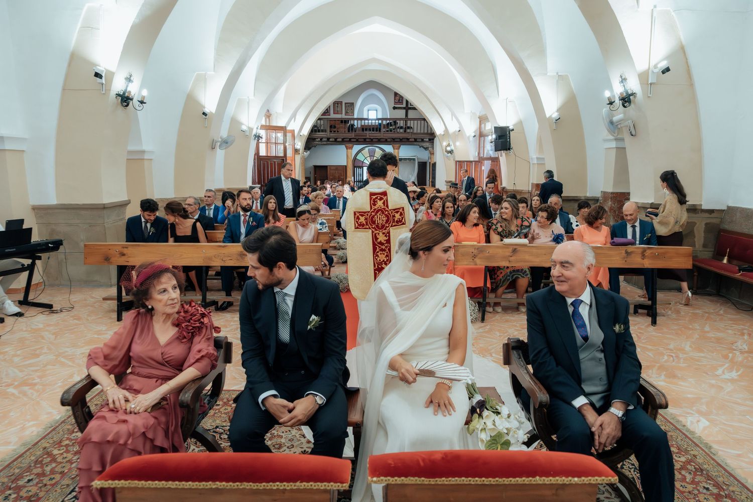 Bride and groom seated at church altar during wedding ceremony with guests filling the white-walled chapel.