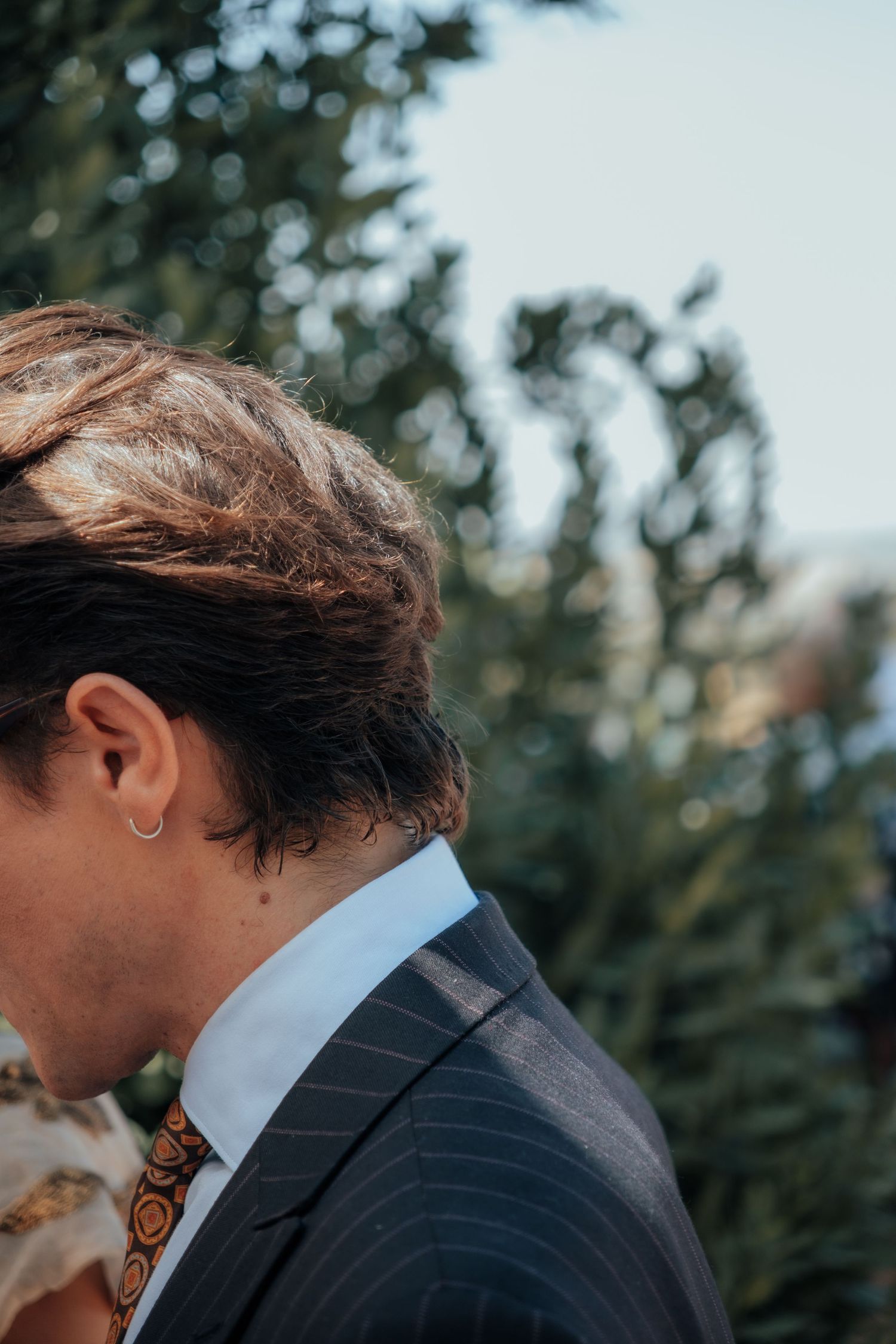 Close-up profile of groom with small ear piercing wearing a pinstripe suit at an outdoor wedding ceremony.