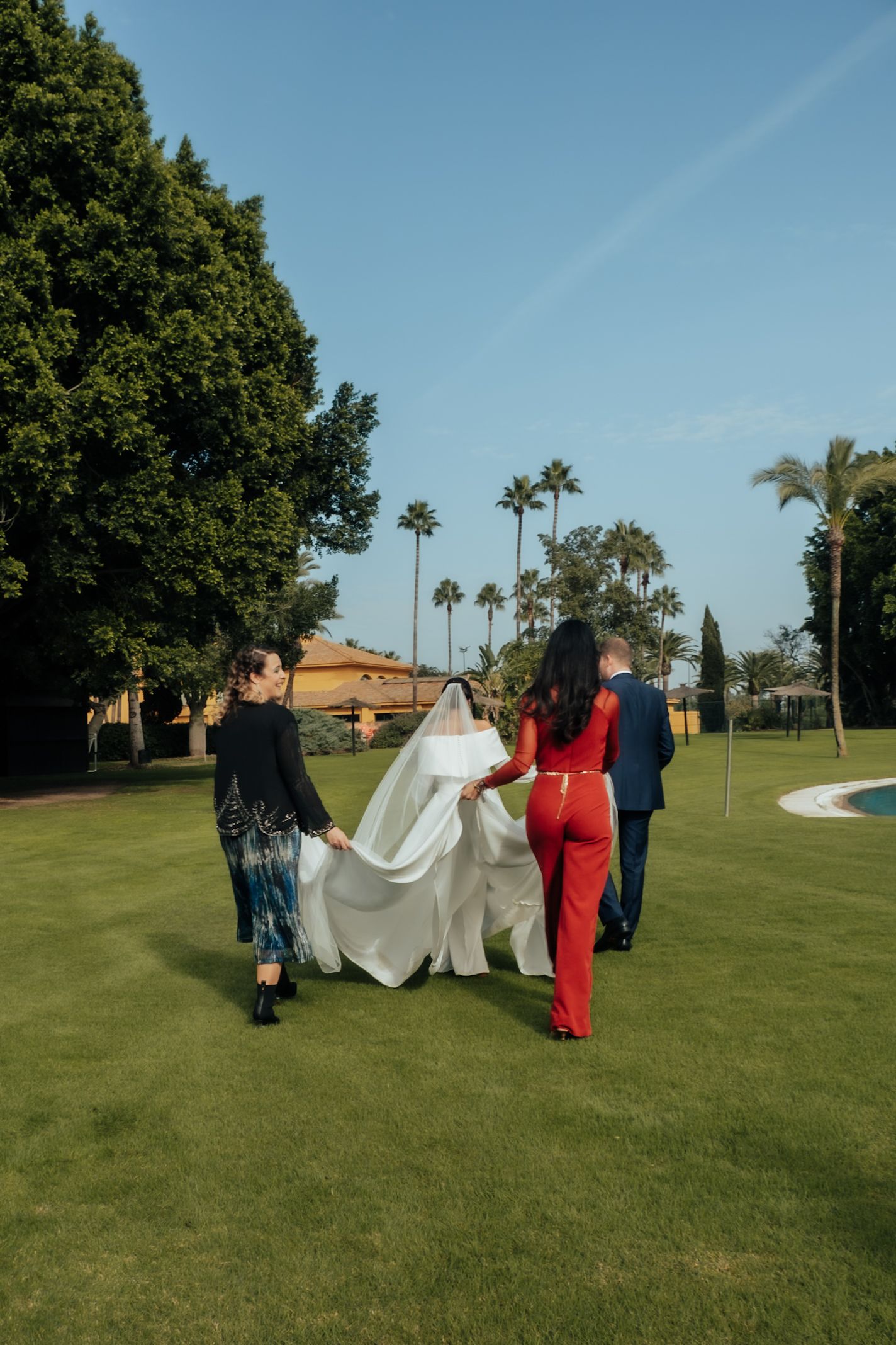 Bride in white gown with long train being assisted by bridesmaids in red and blue outfits on lawn.