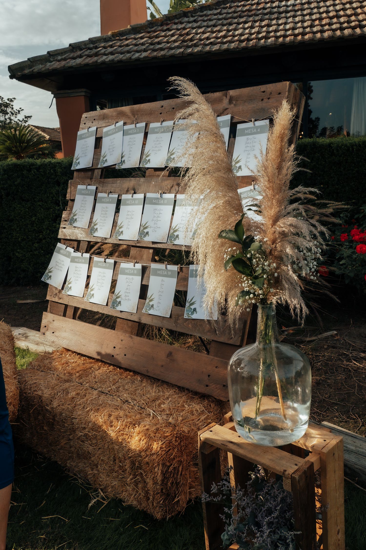 Rustic wooden pallet seating chart display with pampas grass in glass vase on hay bale at wedding.