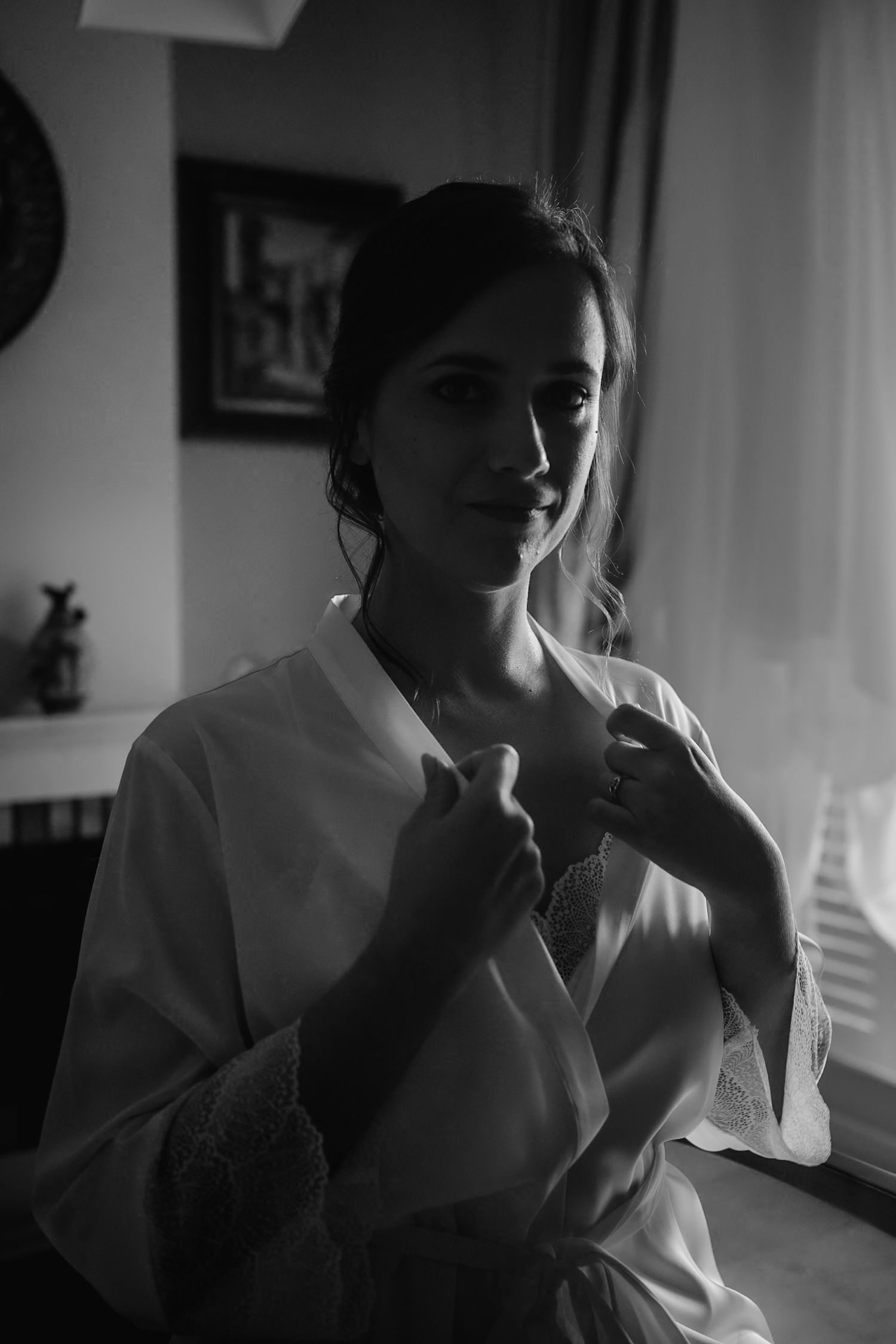 Black and white portrait of a bride in a white lace robe adjusting her neckline before her wedding.