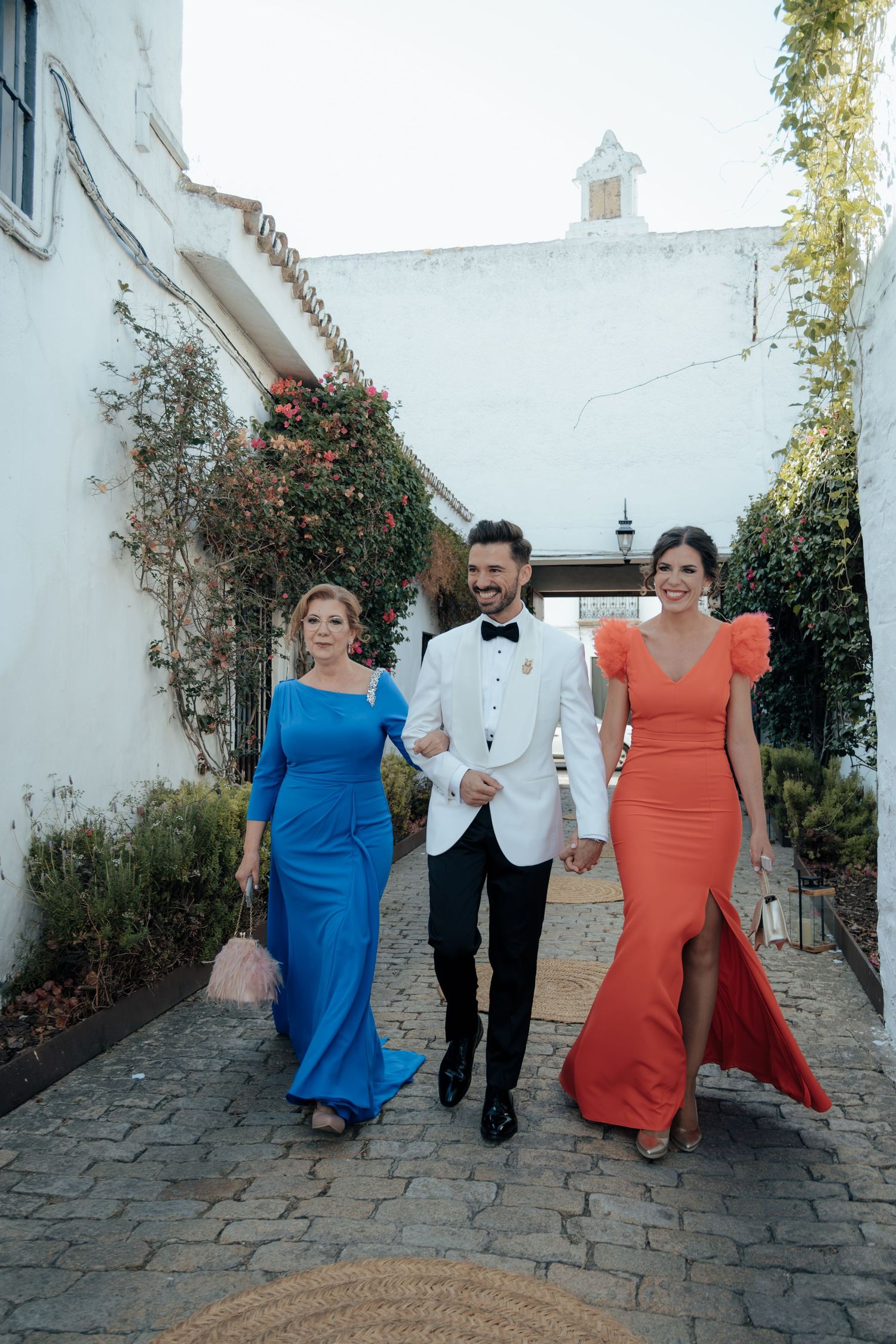 Groom in white tuxedo walks with two women in blue and orange gowns at outdoor wedding venue.