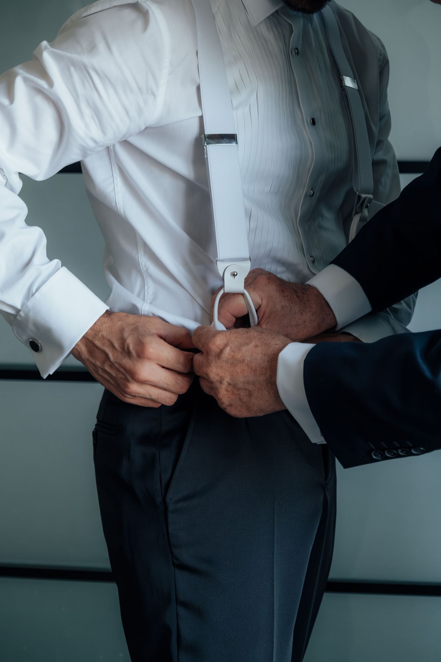Close-up of hands fastening suspenders onto a white tuxedo shirt during wedding preparations.
