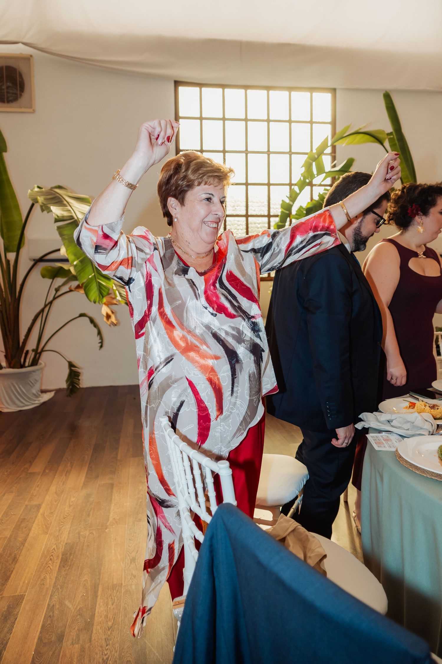 Elderly woman in colorful patterned dress dancing joyfully with arms raised at a wedding reception.