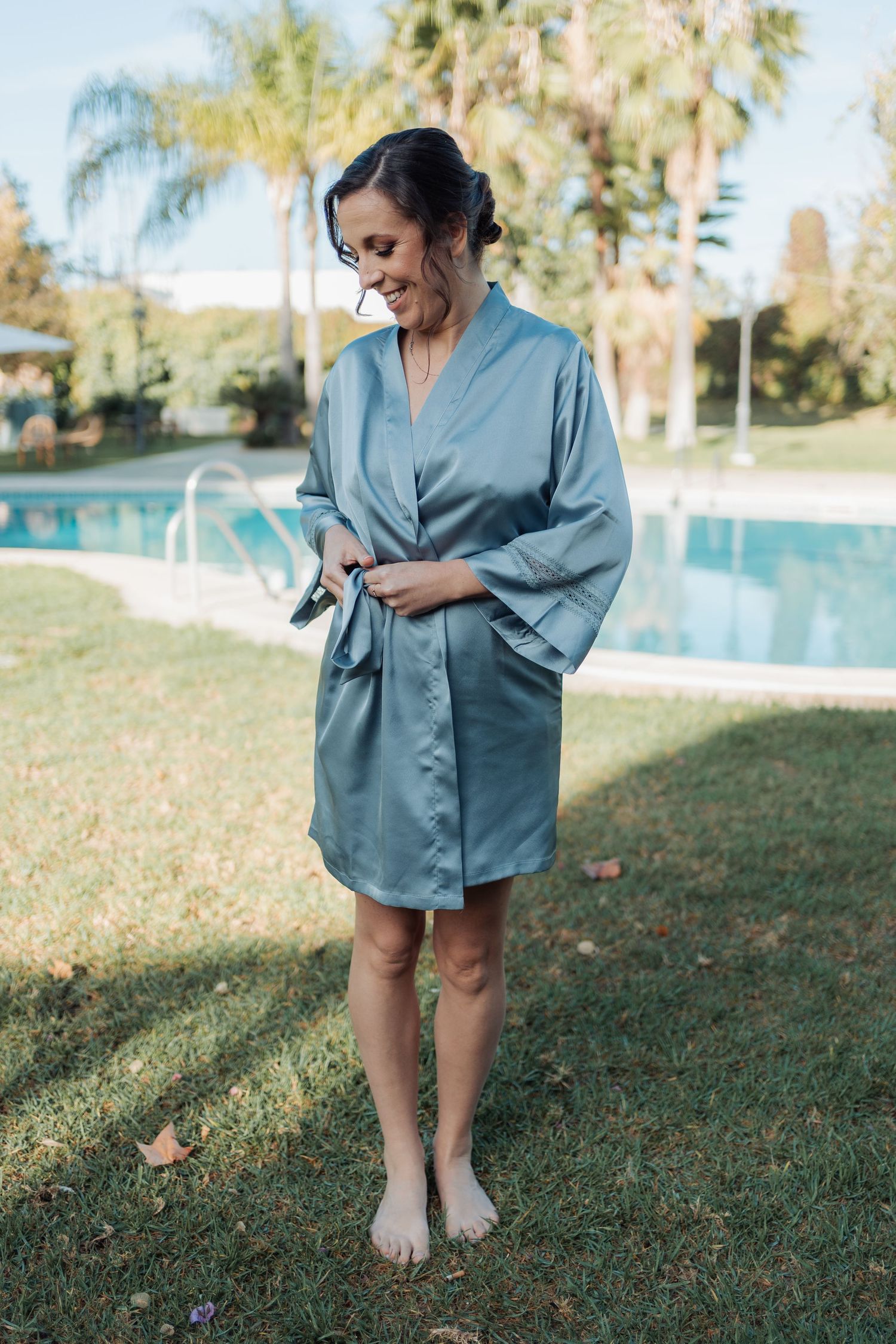 Bride in teal satin robe standing barefoot on grass beside outdoor swimming pool.