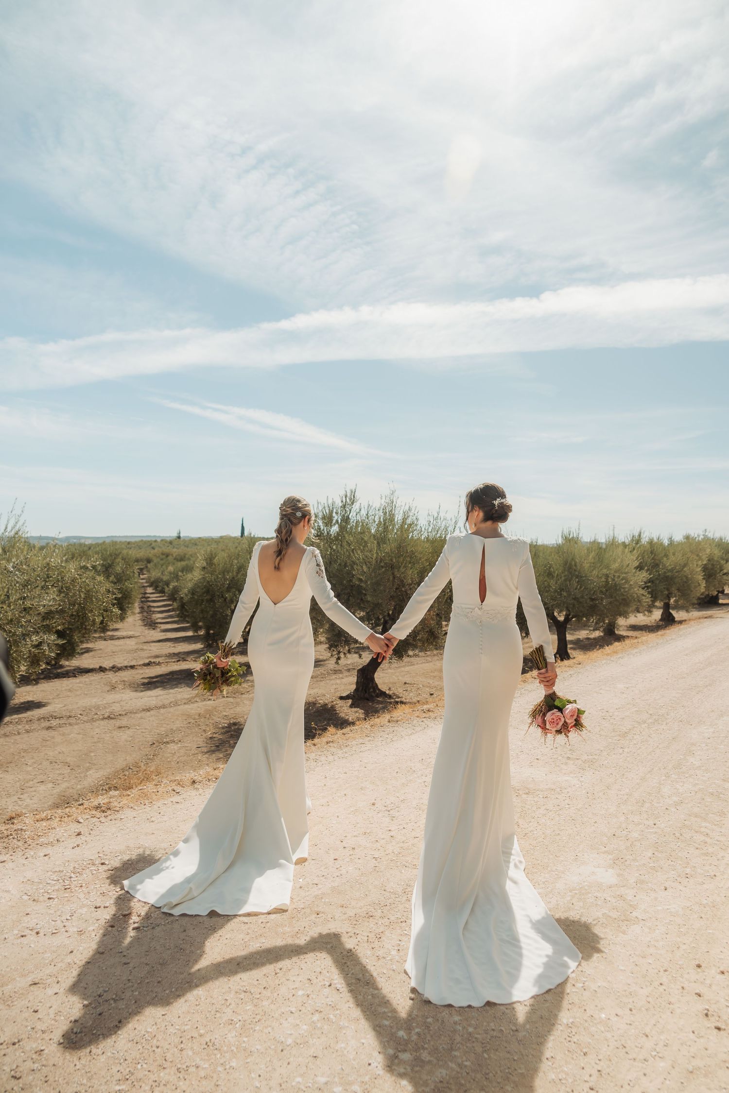Two brides in white gowns holding hands walking down a sandy path through an olive grove.