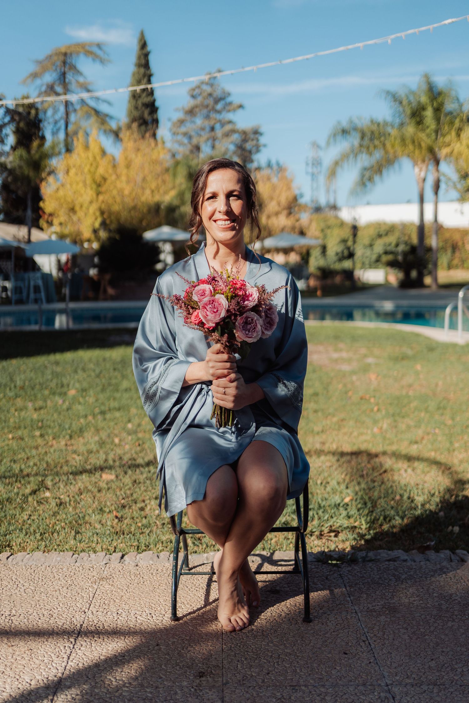 Smiling bride in blue robe sits barefoot on chair holding pink bouquet in sunny garden near pool.