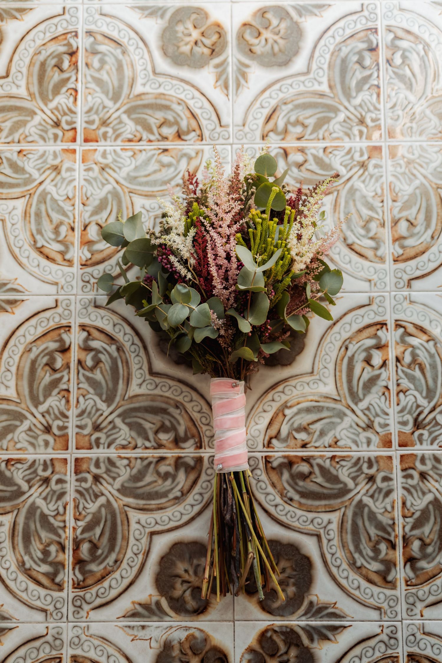 Rustic bridal bouquet with eucalyptus and pink wildflowers tied with ribbon against ornate vintage tiles.