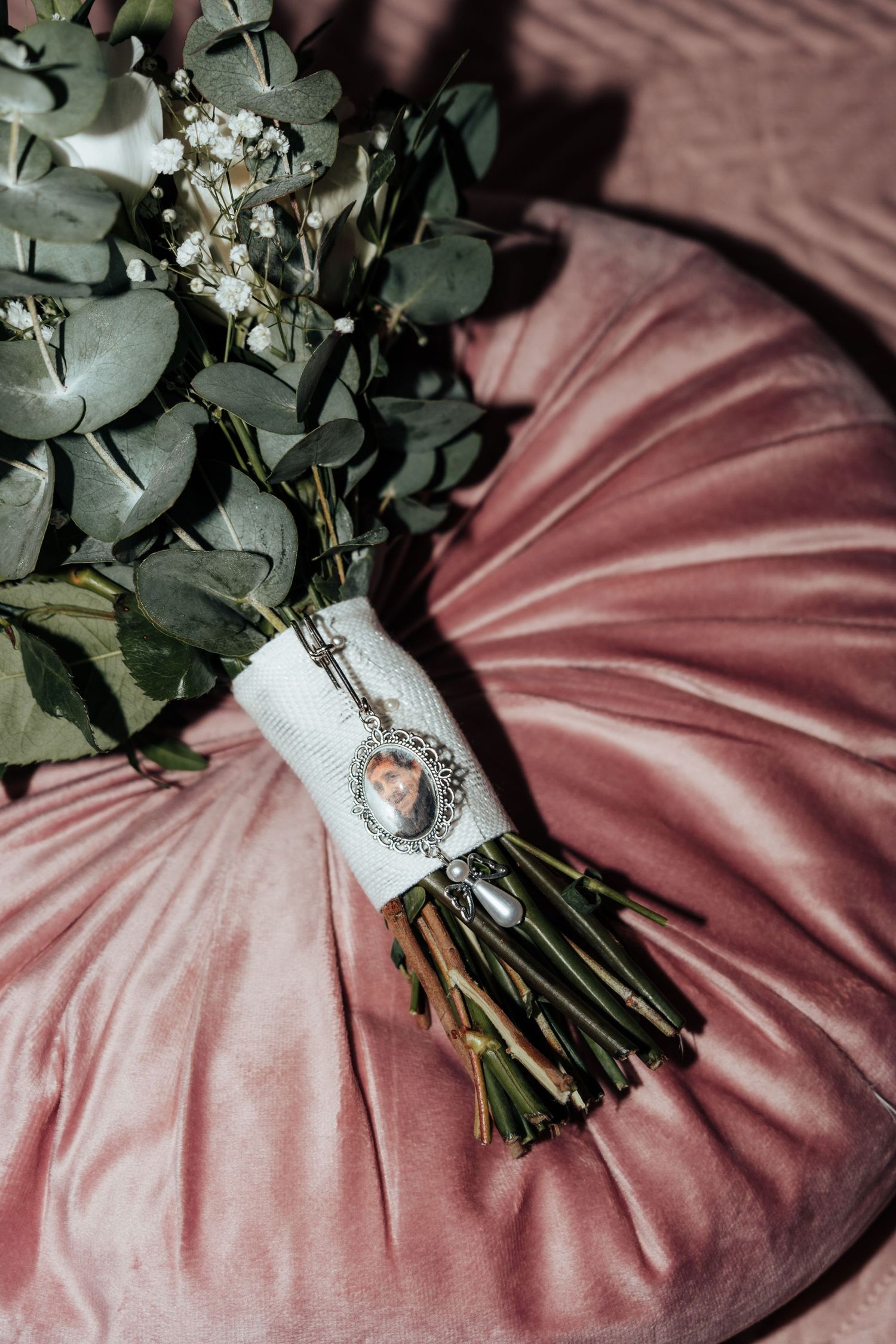 Bridal bouquet with eucalyptus and baby's breath tied with white ribbon and locket charm on pink velvet pillow.