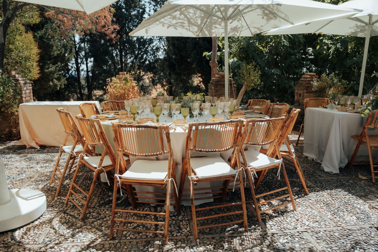 Outdoor wedding reception with round tables, bamboo chairs, white umbrellas, and green glassware on a gravel patio.