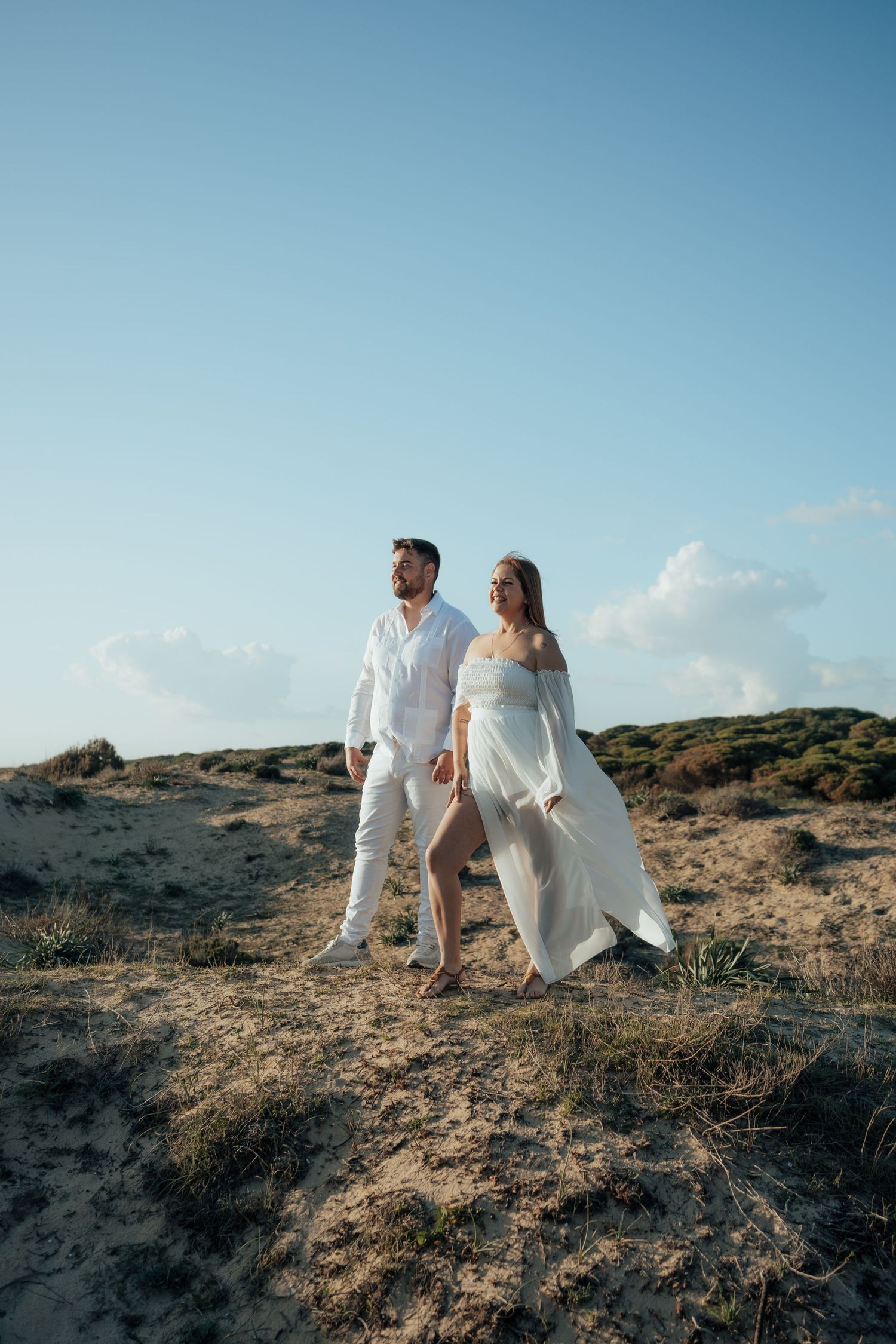 Couple in white outfits walking on sandy dunes under a clear blue sky, perfect for beach wedding inspiration.
