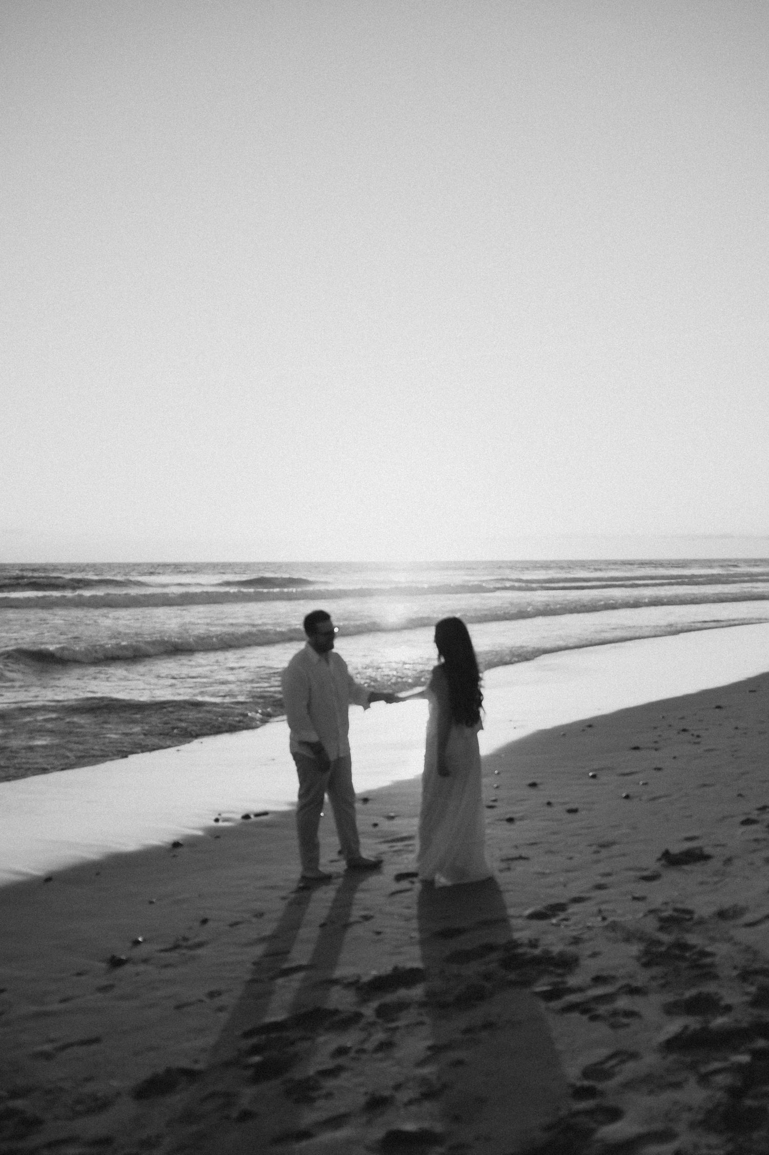 Black and white photo of a couple holding hands on a sandy beach at sunset with ocean waves in the background.