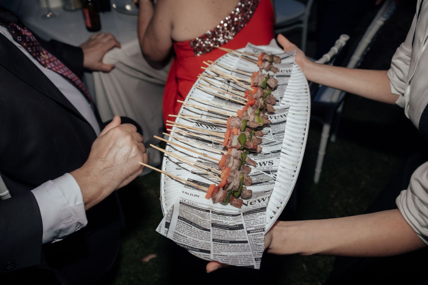 Catering server presenting skewered appetizers on a newspaper-lined tray at an outdoor party.