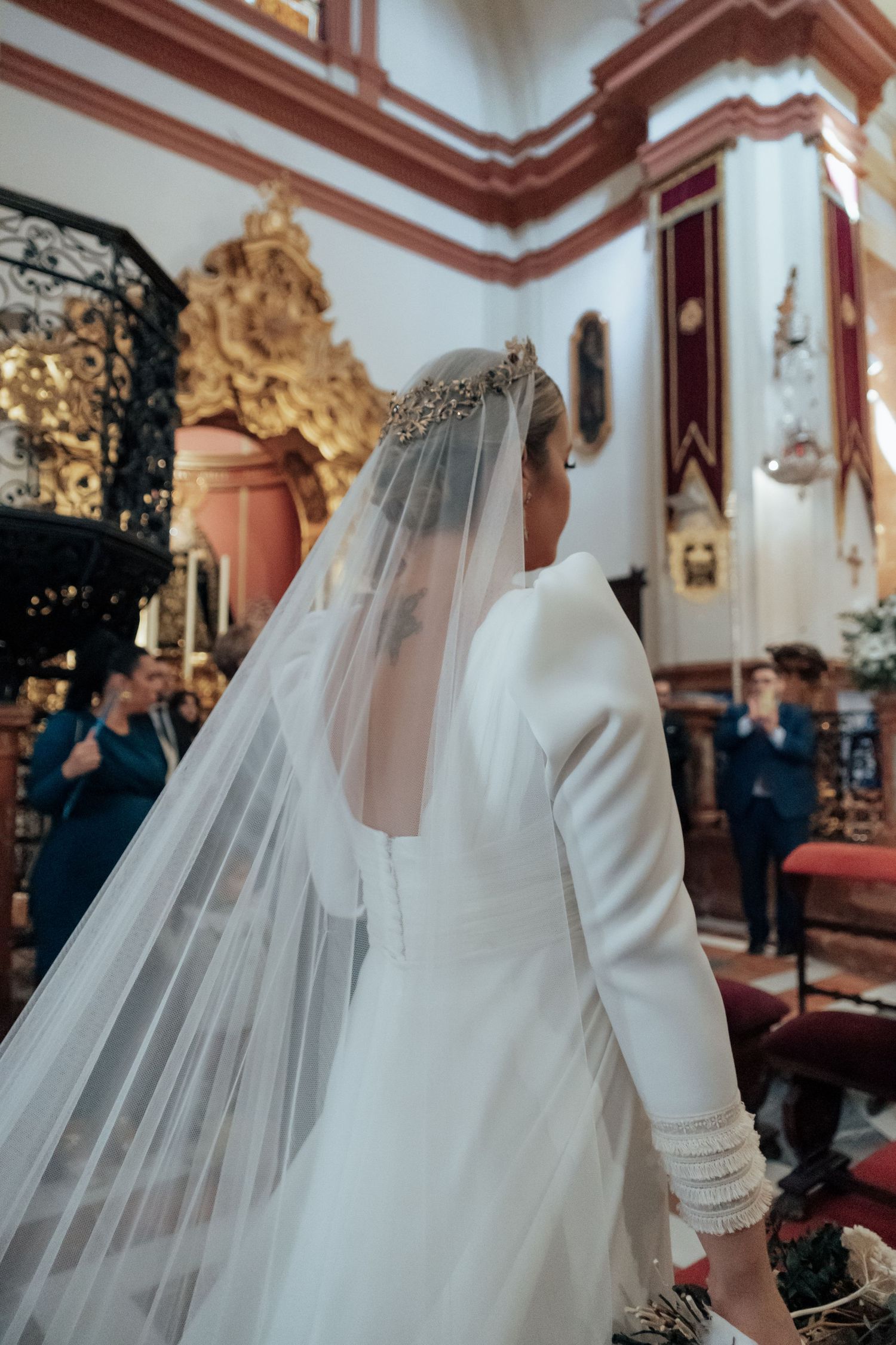 Bride in white gown with gold crown and cathedral veil inside ornate baroque church.