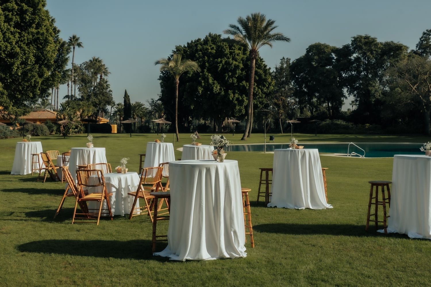 Elegant outdoor wedding cocktail reception setup with white-draped round tables on a green lawn beside a pool with palm trees.