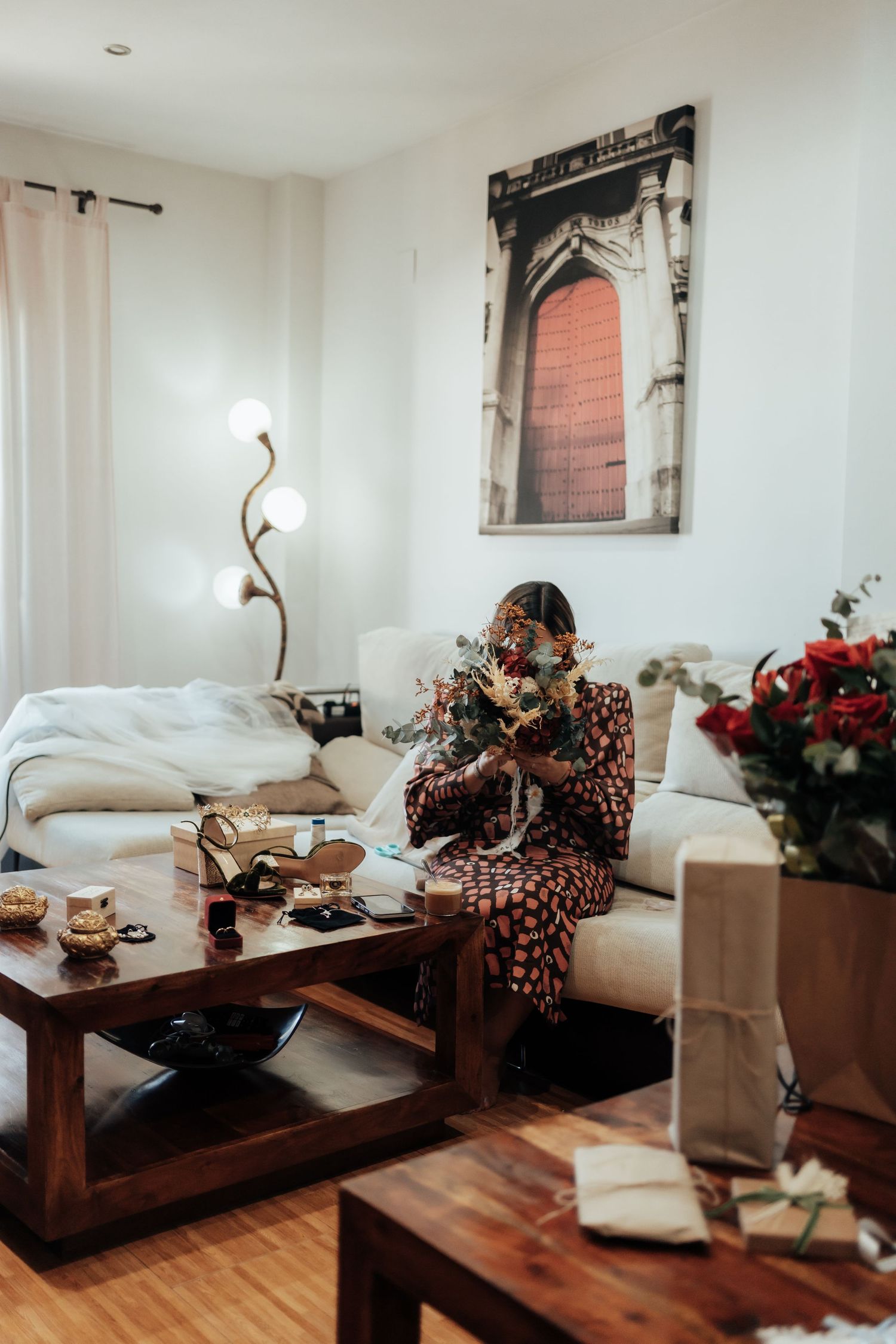 Bride in floral dress arranging bouquet on sofa in bright modern living room before wedding.