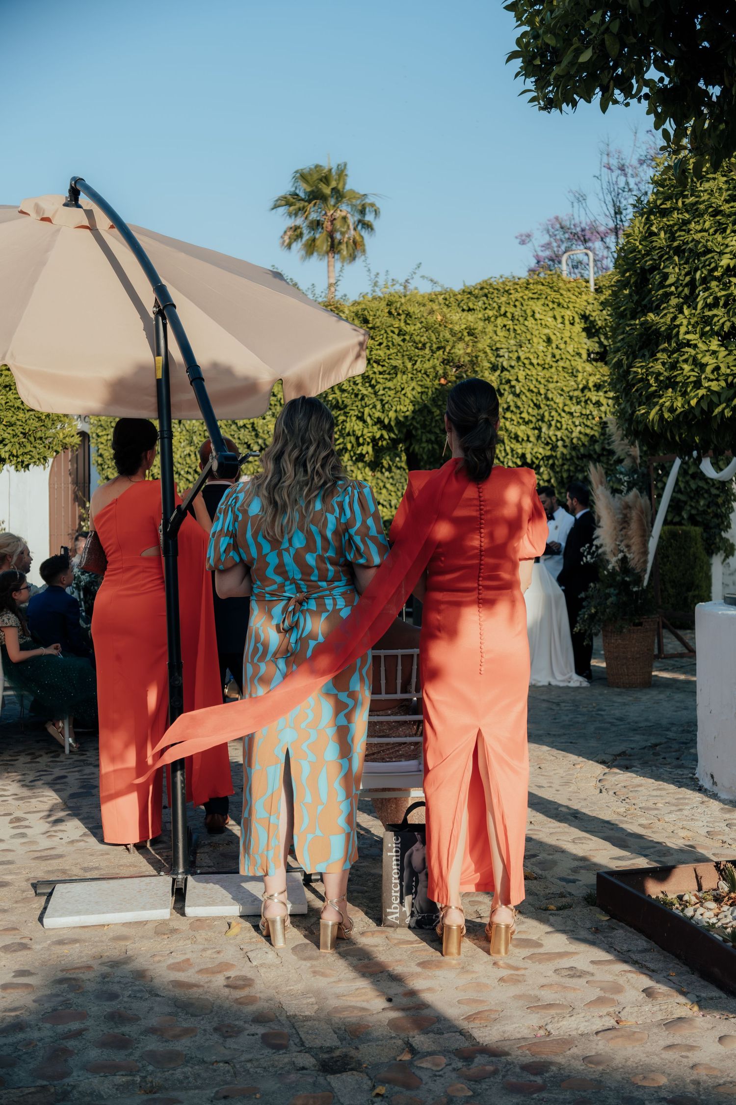Three women in colorful formal attire stand watching outdoor wedding ceremony under large umbrella.