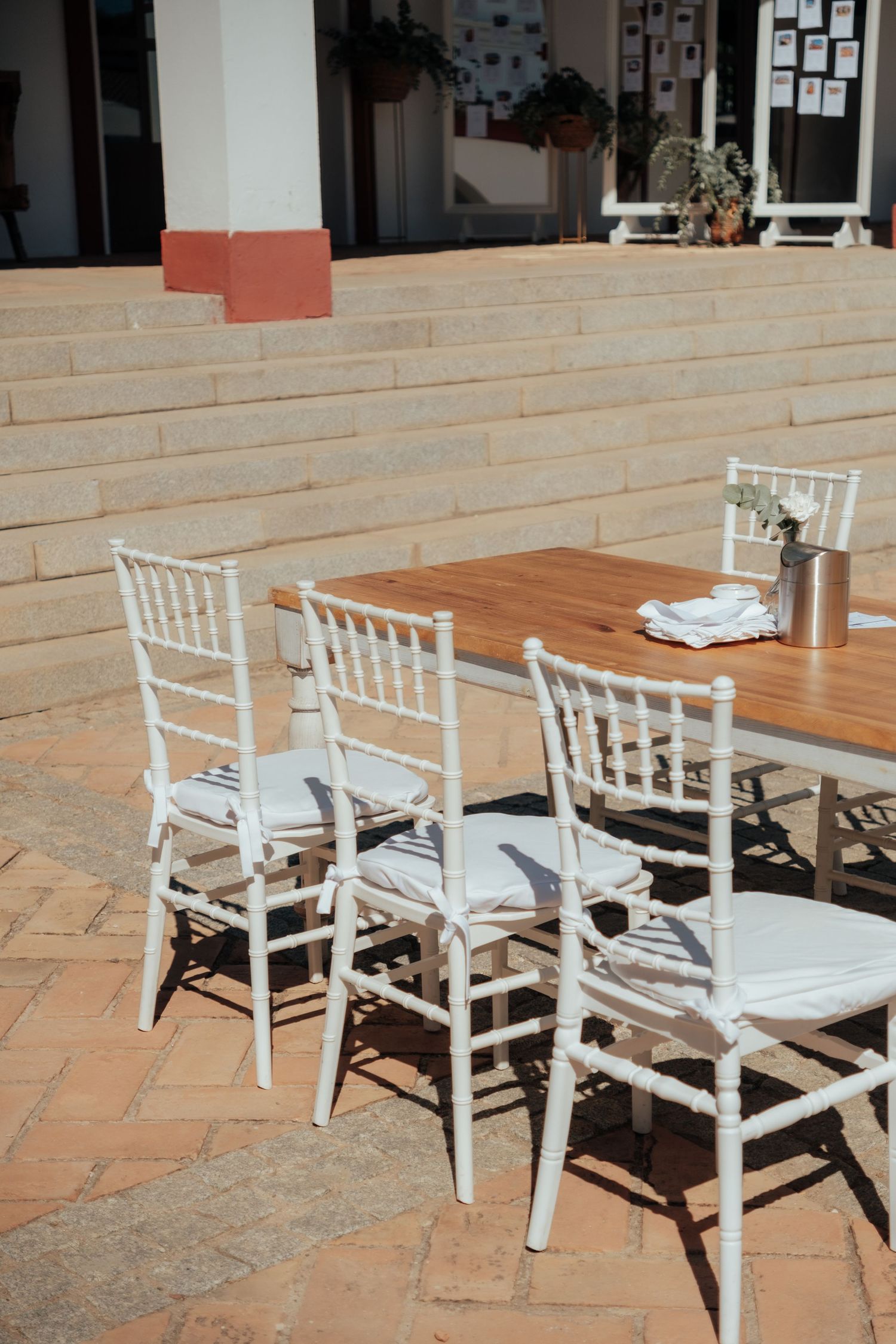 White Chiavari chairs and wooden table set up outdoors on brick patio in sunlight.