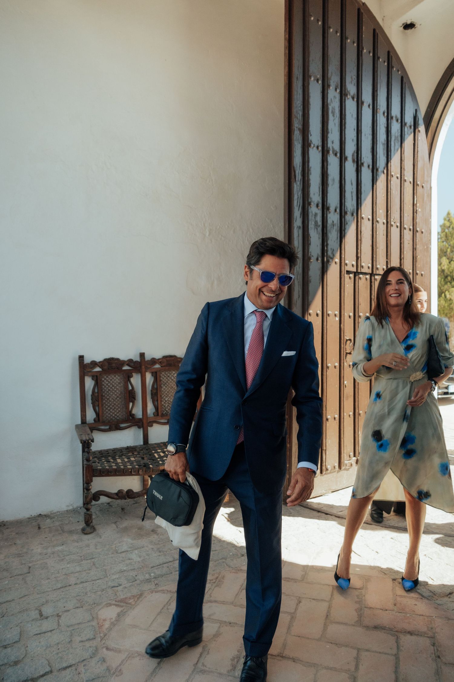 Smiling groom in navy suit with sunglasses walks through hacienda doorway as woman laughs behind him.