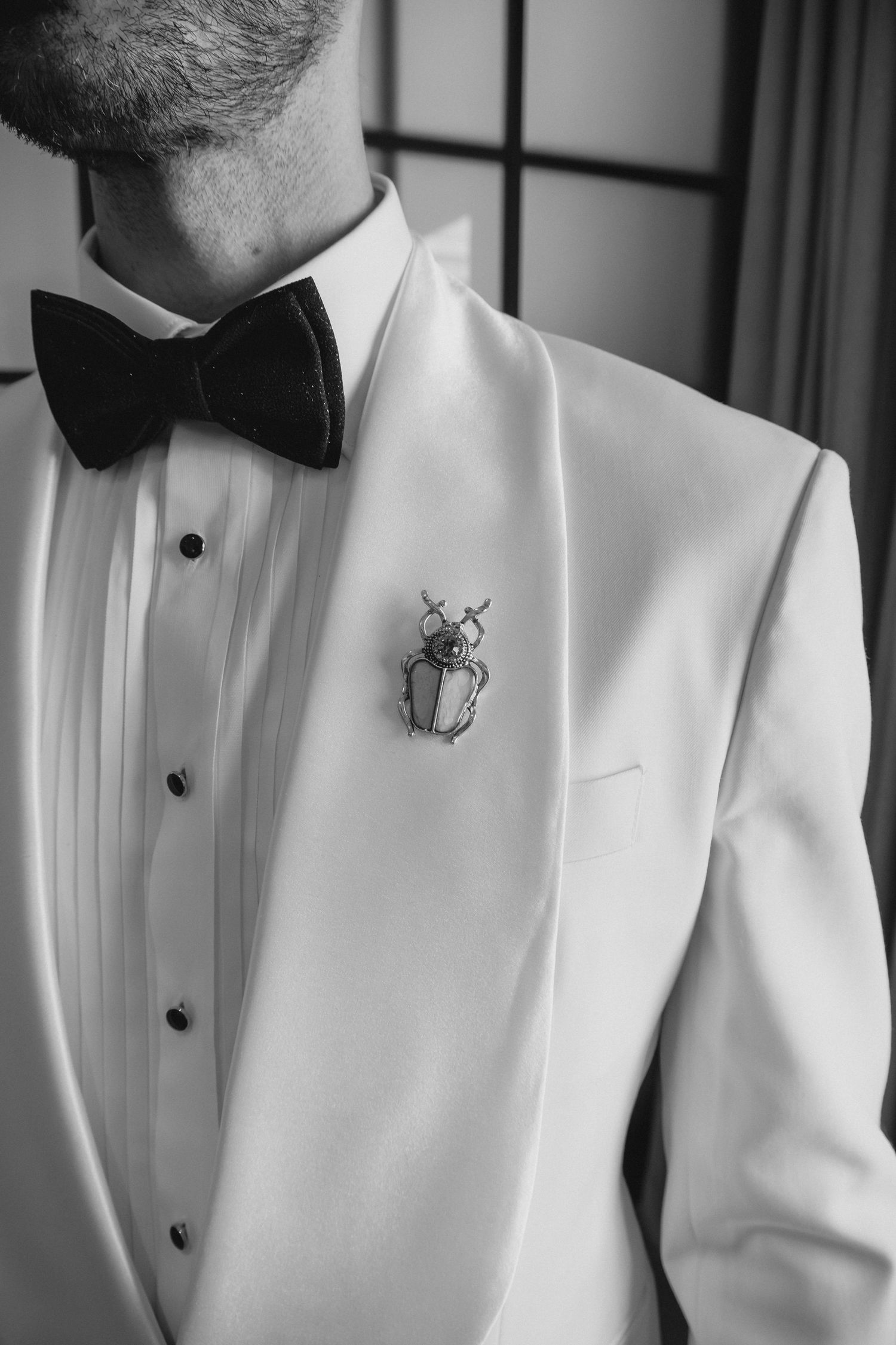 Close-up black and white shot of a groom's white tuxedo lapel with bow tie and beetle brooch.
