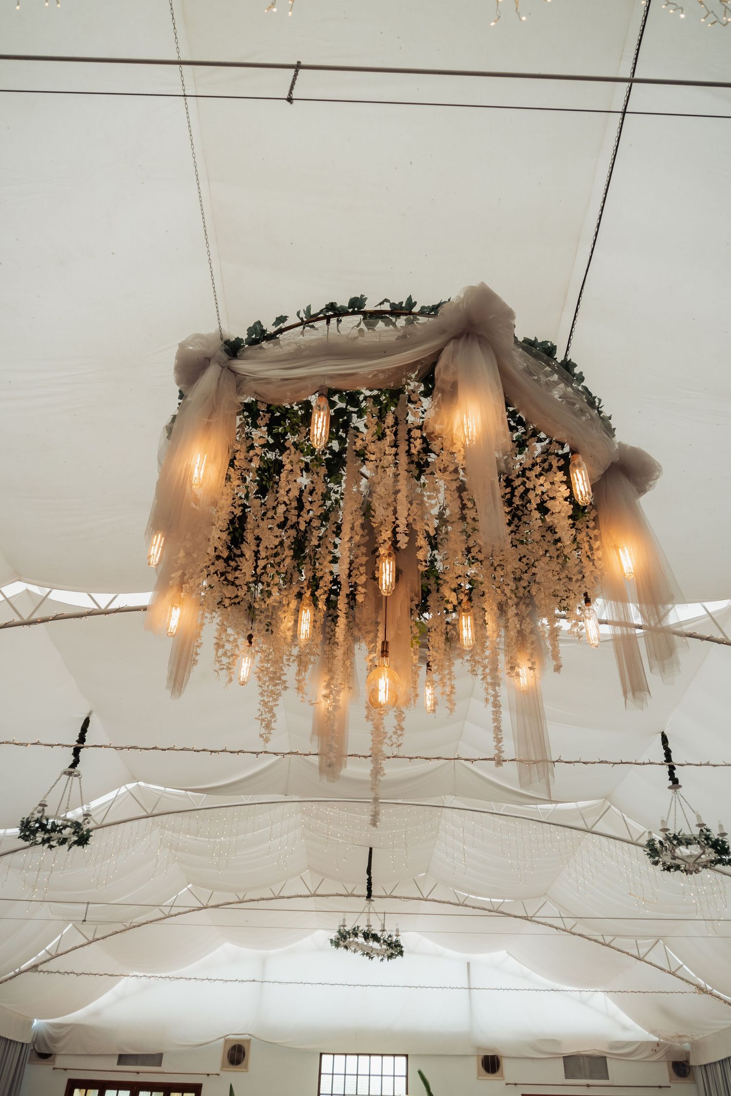 Ornate wedding chandelier with Edison bulbs, flowing fabric, and cascading greenery under white tent.