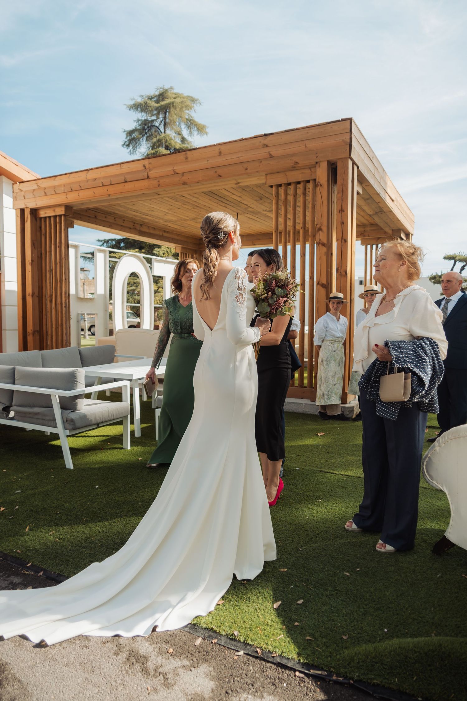 Bride in a long white wedding dress with train stands outdoors near a wooden pergola with guests.