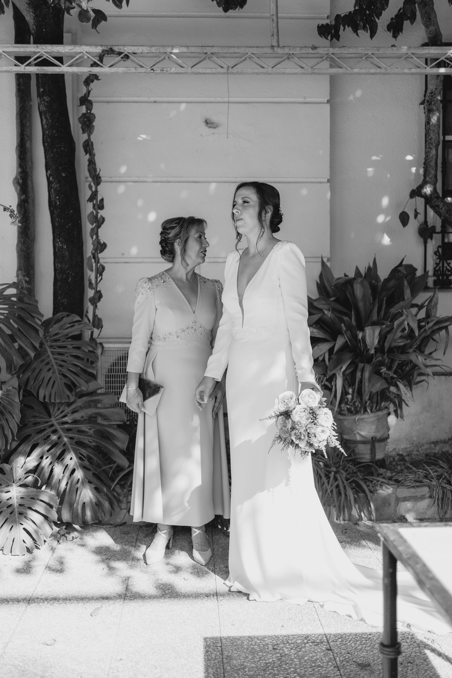Black and white photo of a bride in white gown holding bouquet standing with older woman in garden setting.