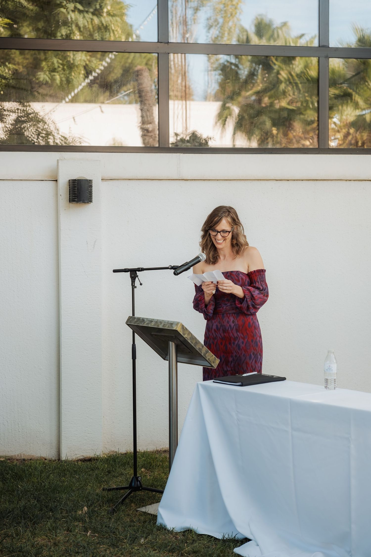 A woman in a red floral off-shoulder dress reads a speech at a podium during an outdoor wedding ceremony.