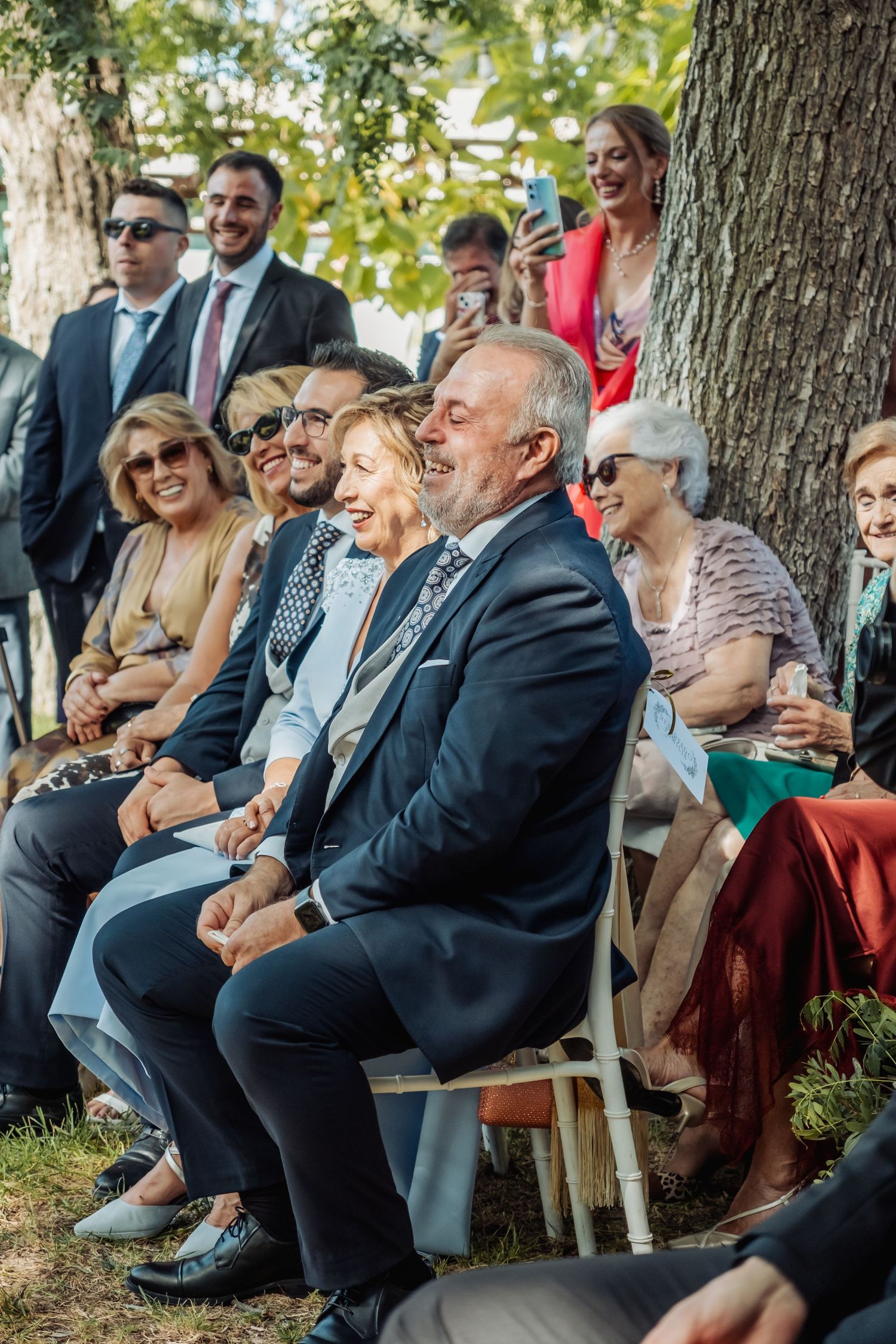 Smiling wedding guests seated outdoors under trees during a sunny garden wedding ceremony.