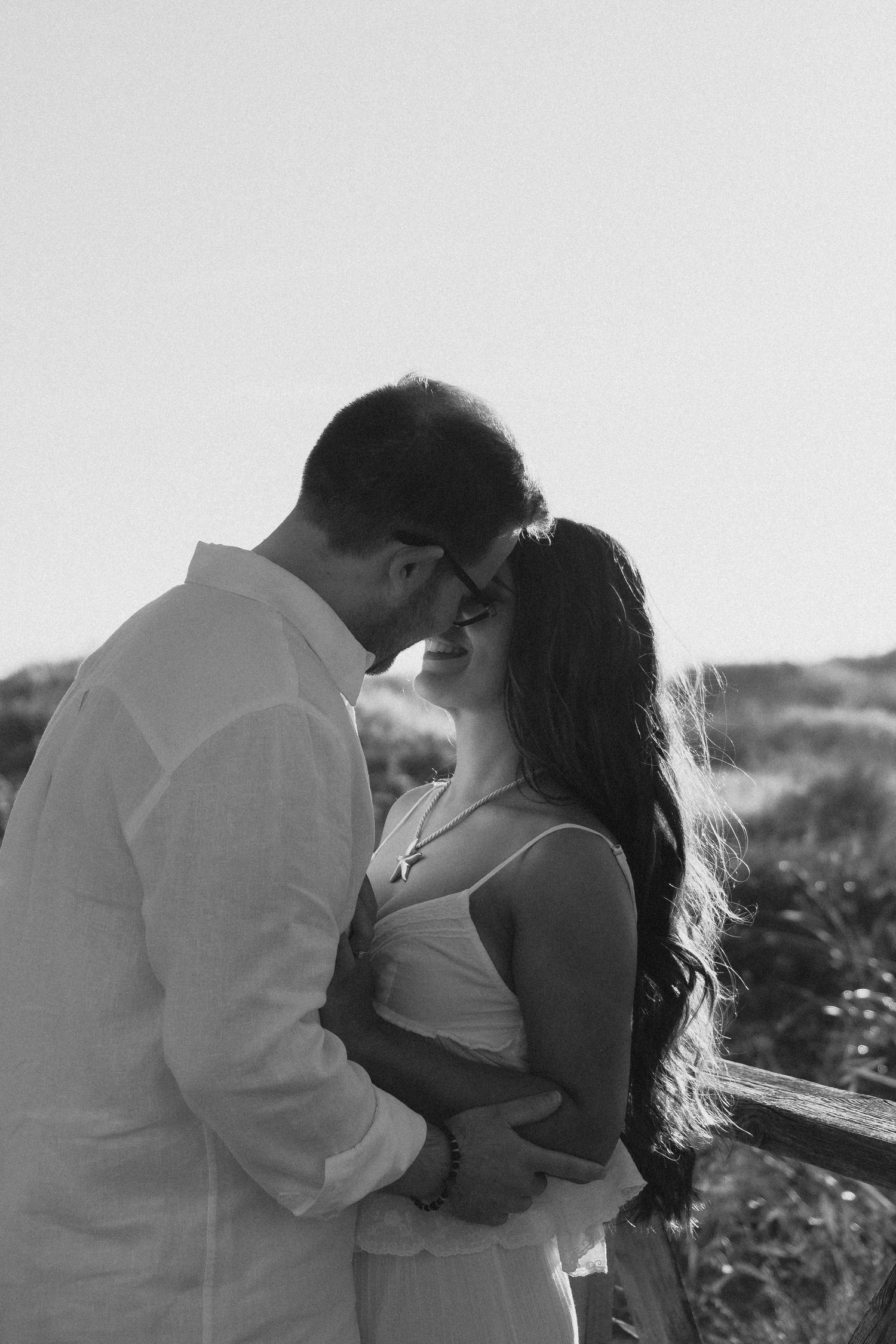 Black and white photo of a romantic couple almost kissing outdoors near a wooden fence.