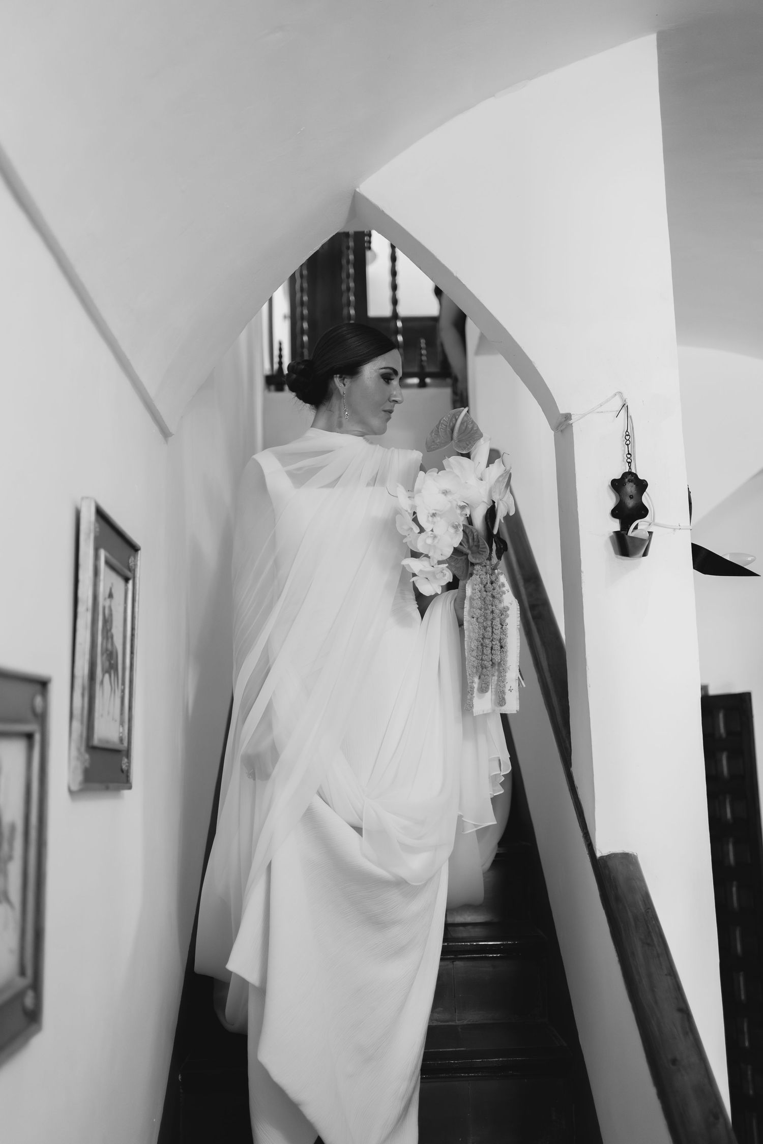 Black and white photo of bride in flowing white dress descending ornate staircase with bouquet.