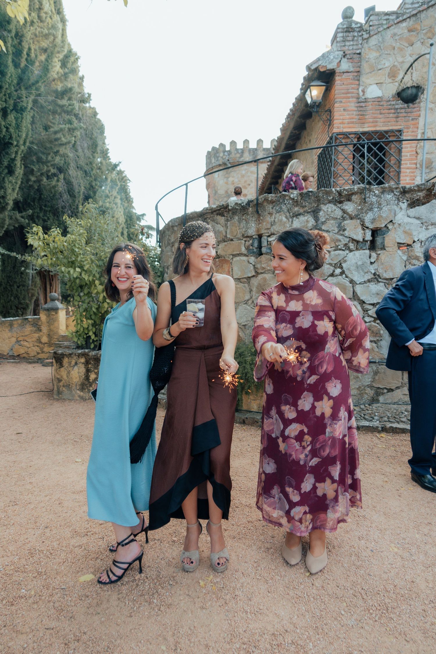 Three female wedding guests in elegant dresses holding sparklers at rustic stone venue.