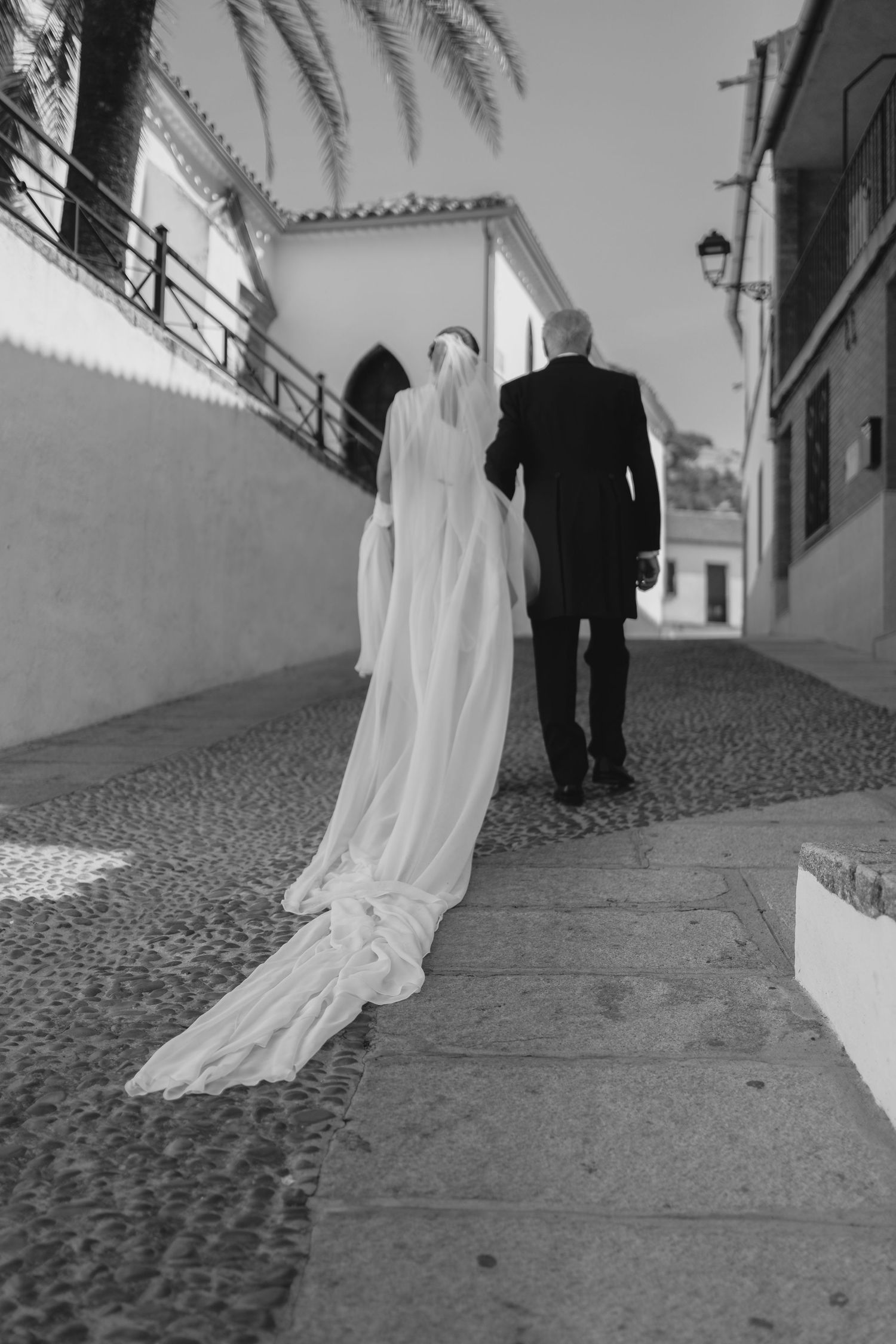 Bride in white gown with long veil walks cobblestone street with older man in black suit.