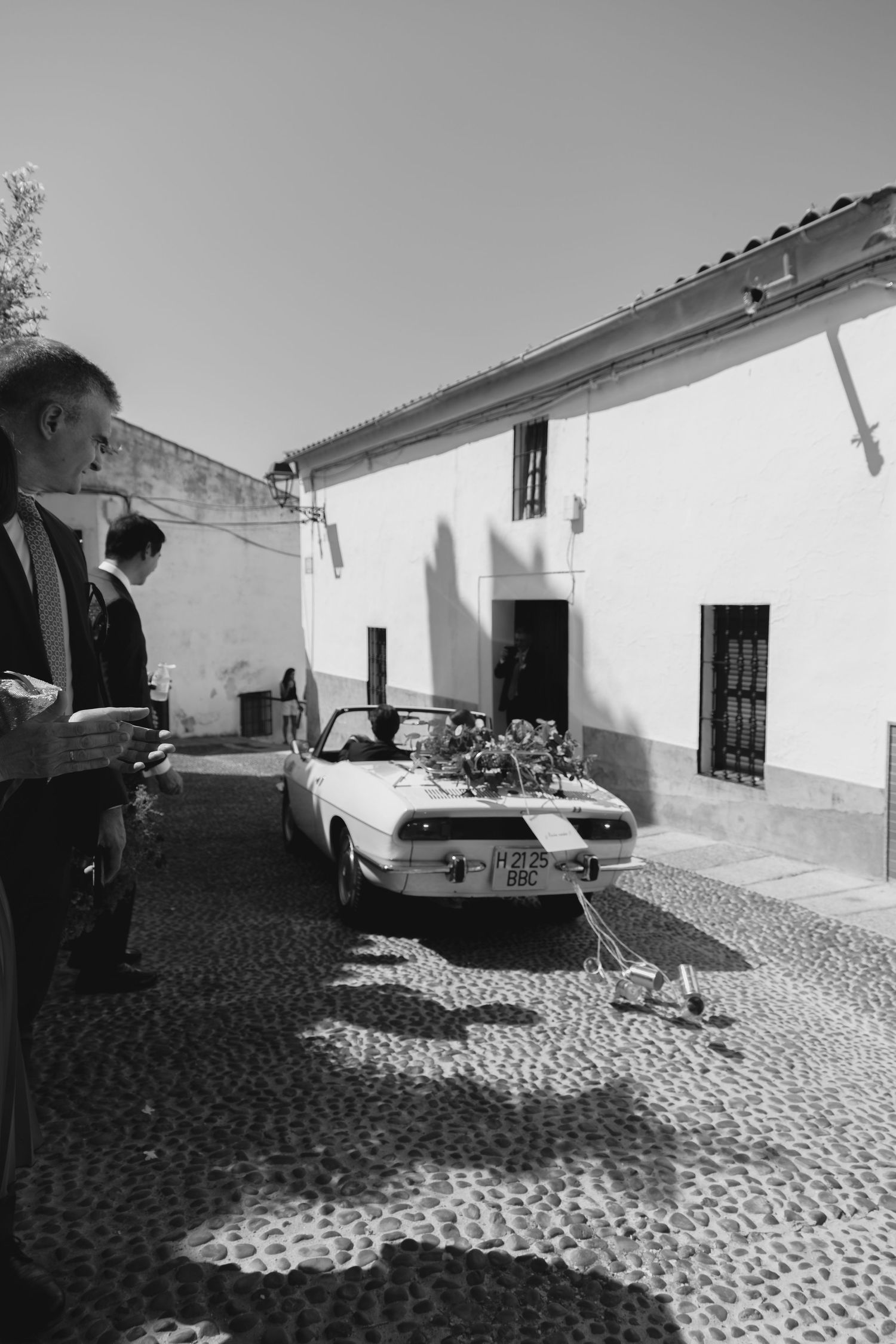 Black and white image of a decorated white vintage convertible car with tin cans on cobblestone street.