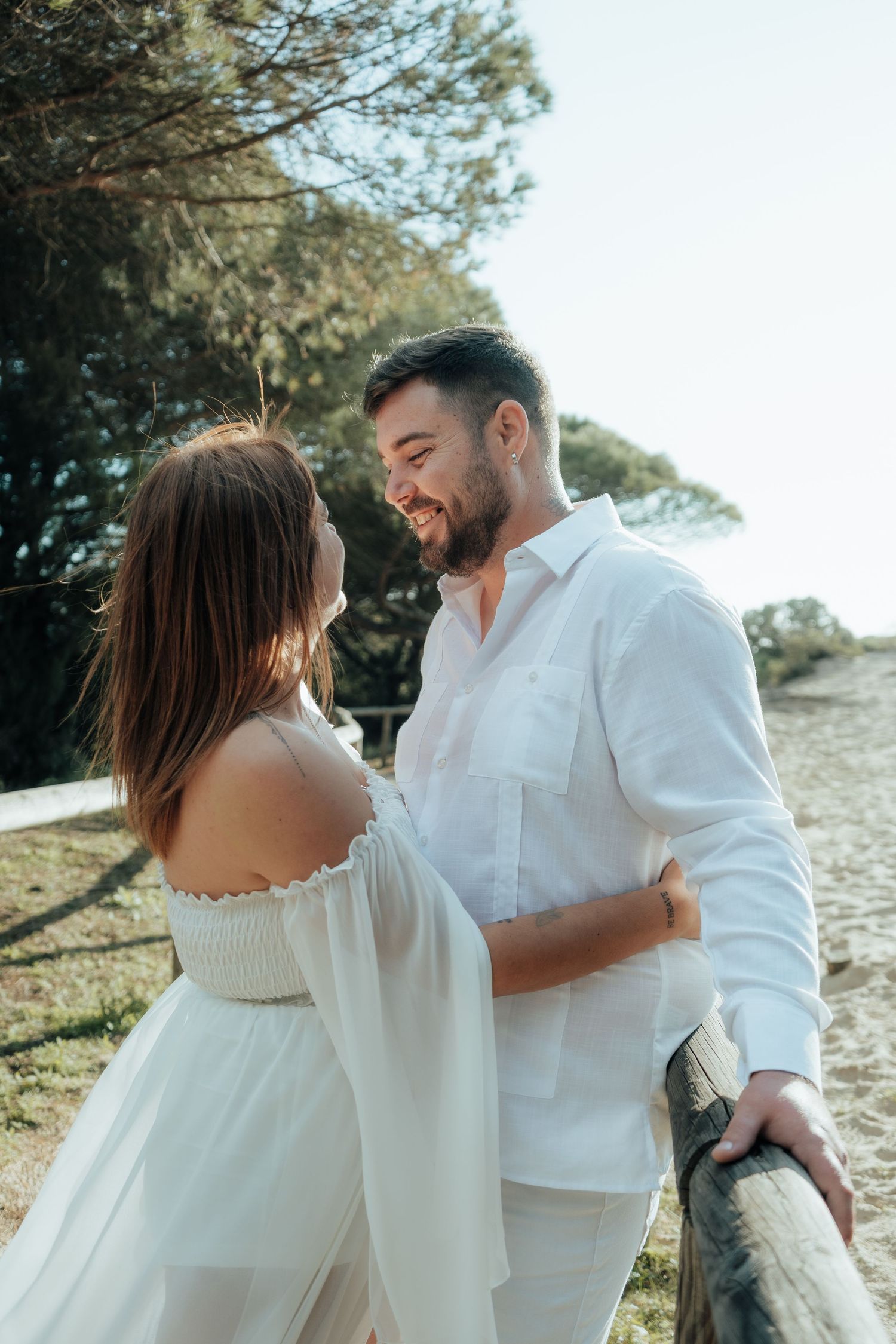 A couple in white outfits share a romantic moment on a sunny beach path surrounded by trees.