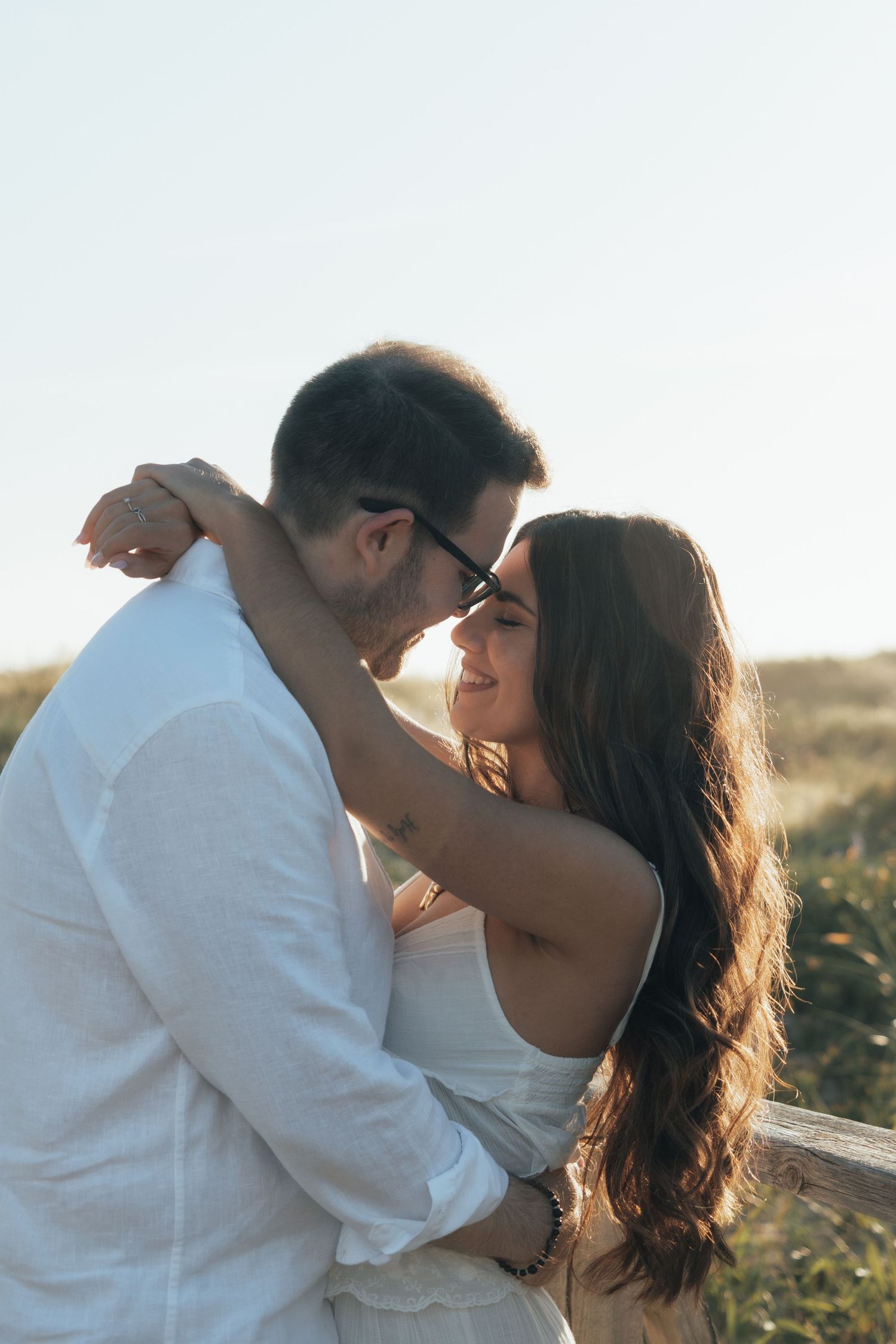 A couple embraces and smiles at each other outdoors in golden sunlight, woman in white dress with long dark hair.