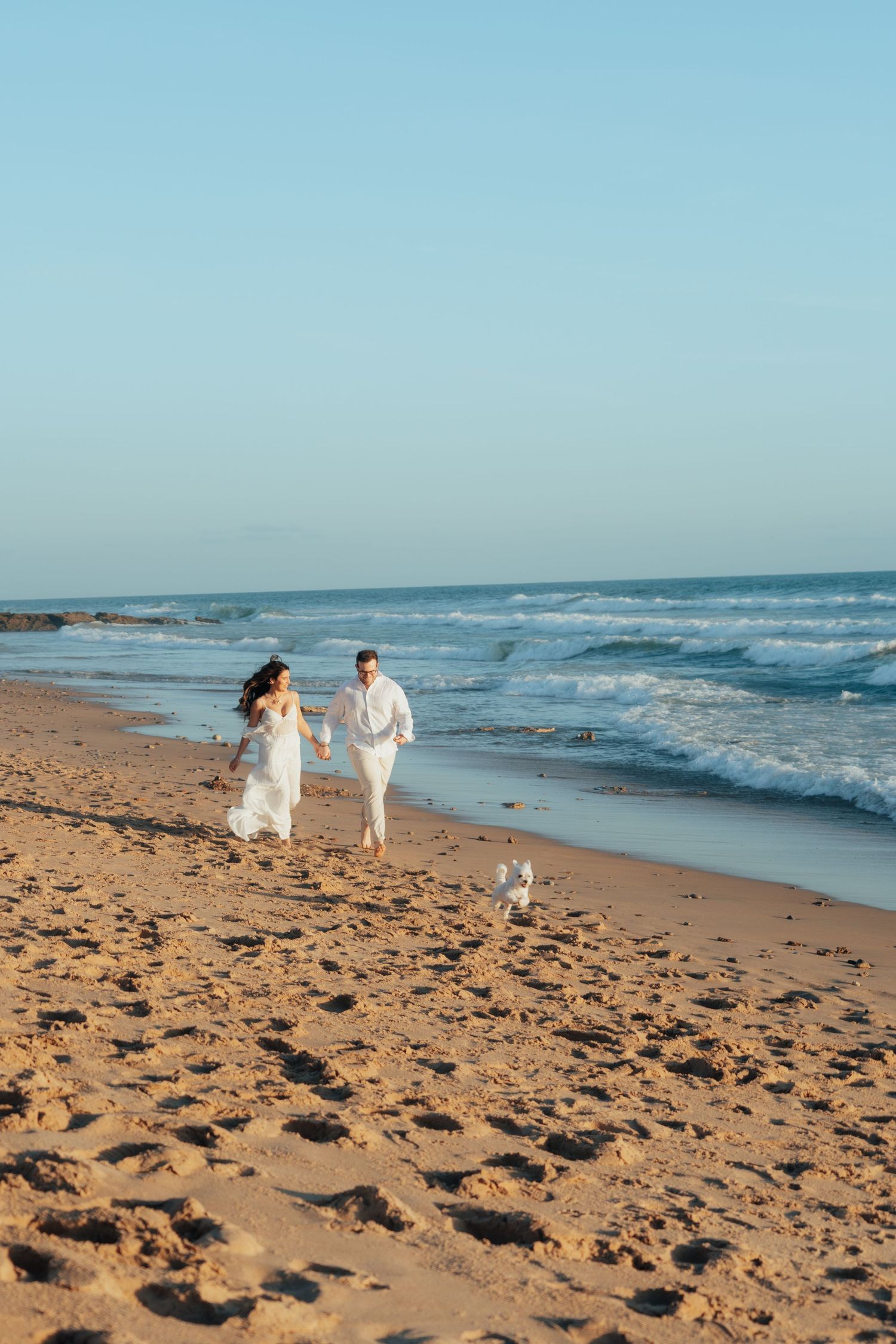 Couple in white attire running joyfully on a sunny beach with a small dog, waves crashing nearby.