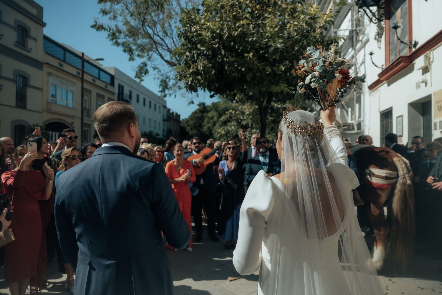 Bride and groom walk through cheering wedding guests outside a sunny urban church after ceremony.