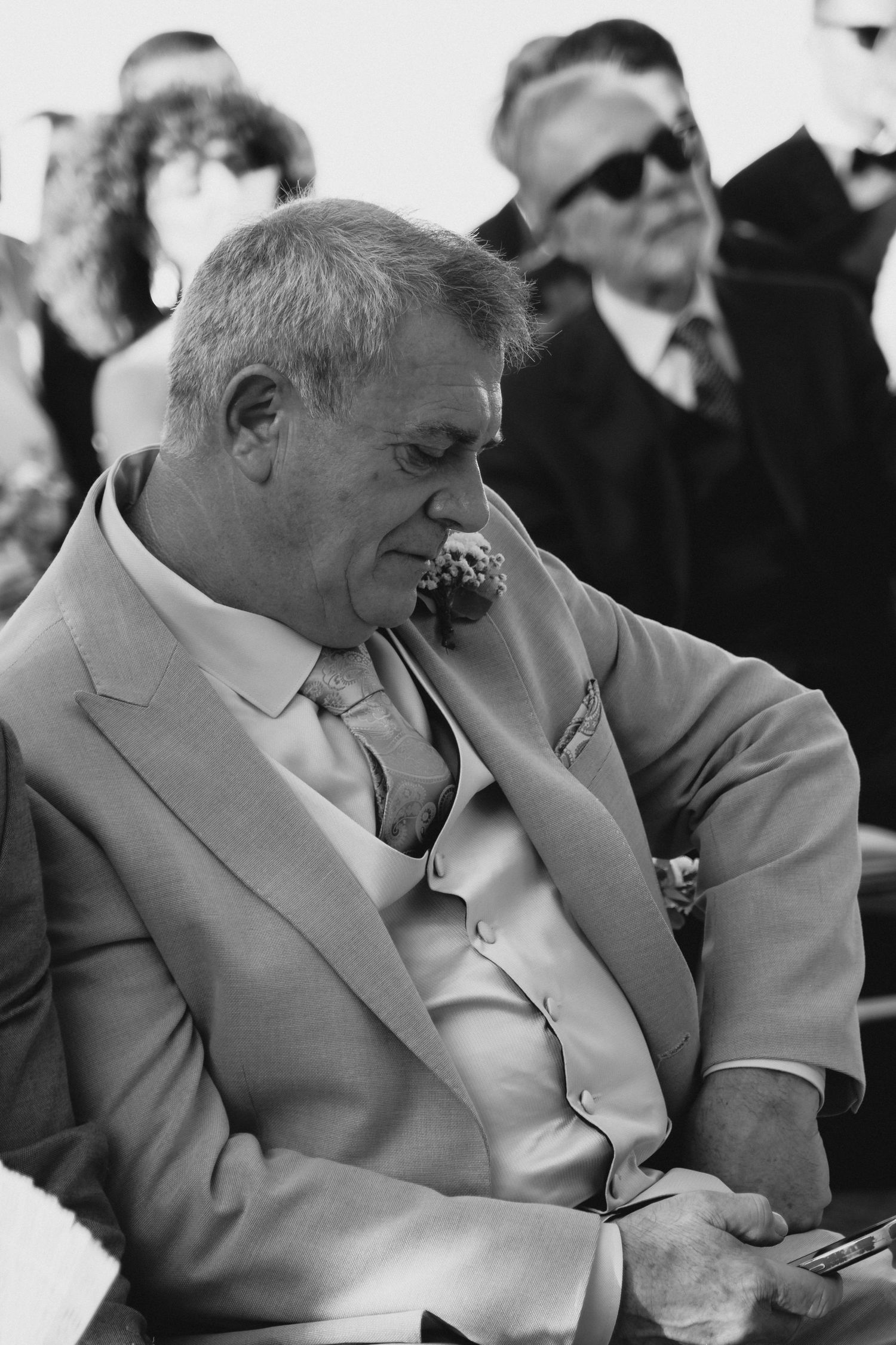 Elderly man in beige suit holds phone during outdoor wedding ceremony in black and white photo.