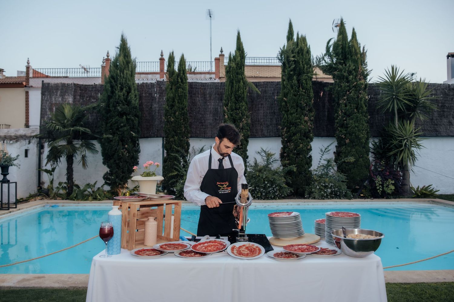 Catering staff member preparing charcuterie display table beside a pool at an outdoor wedding reception.