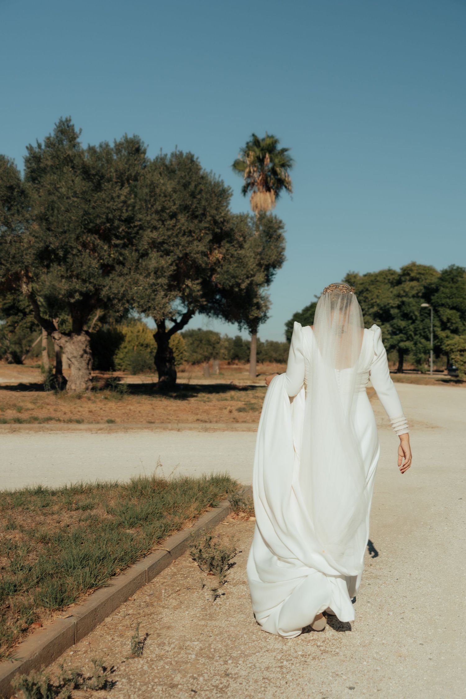 Bride in flowing white gown with veil walking along a sunlit path surrounded by olive trees.