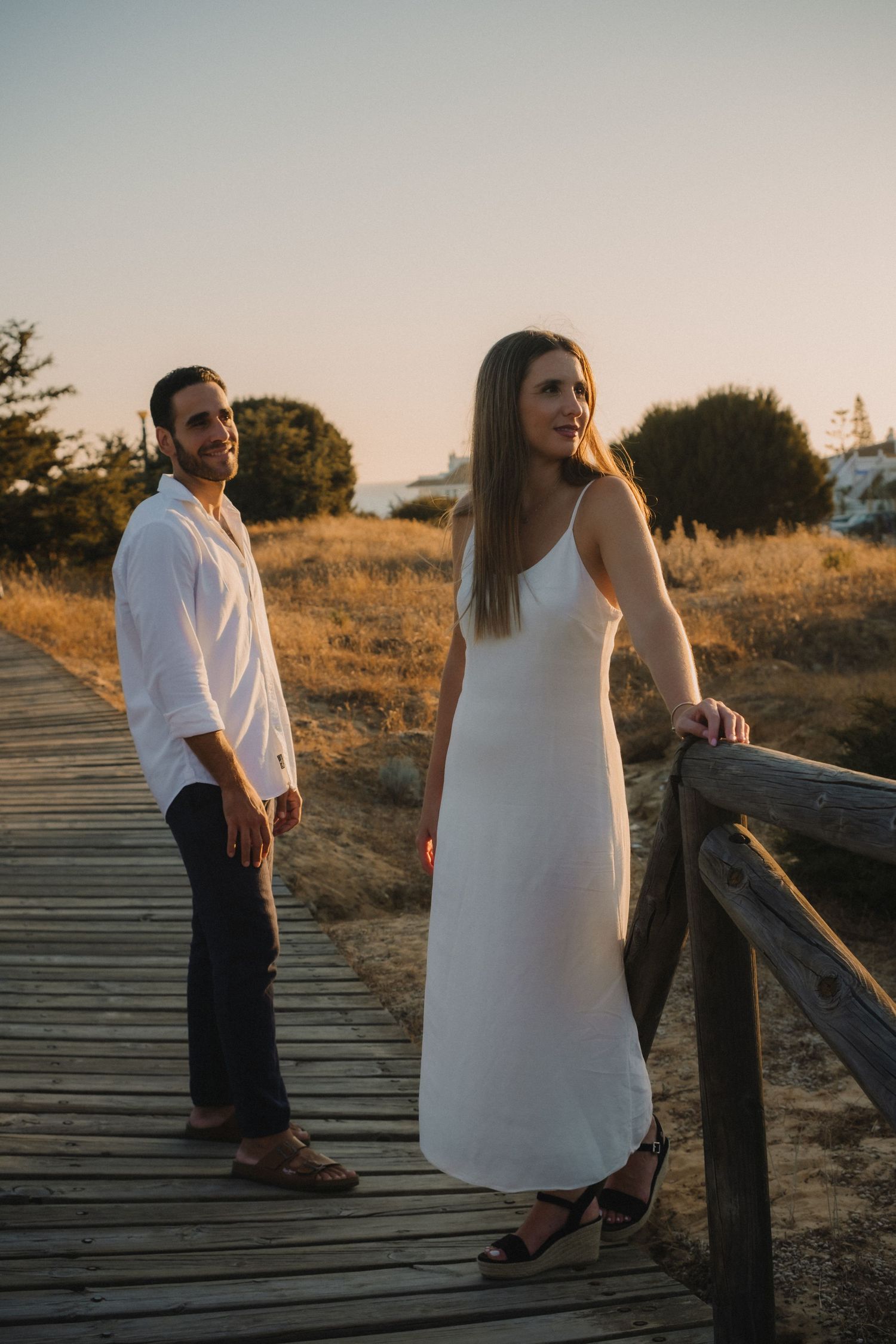 Couple in white outfits standing on a wooden boardwalk at sunset in a golden coastal landscape.