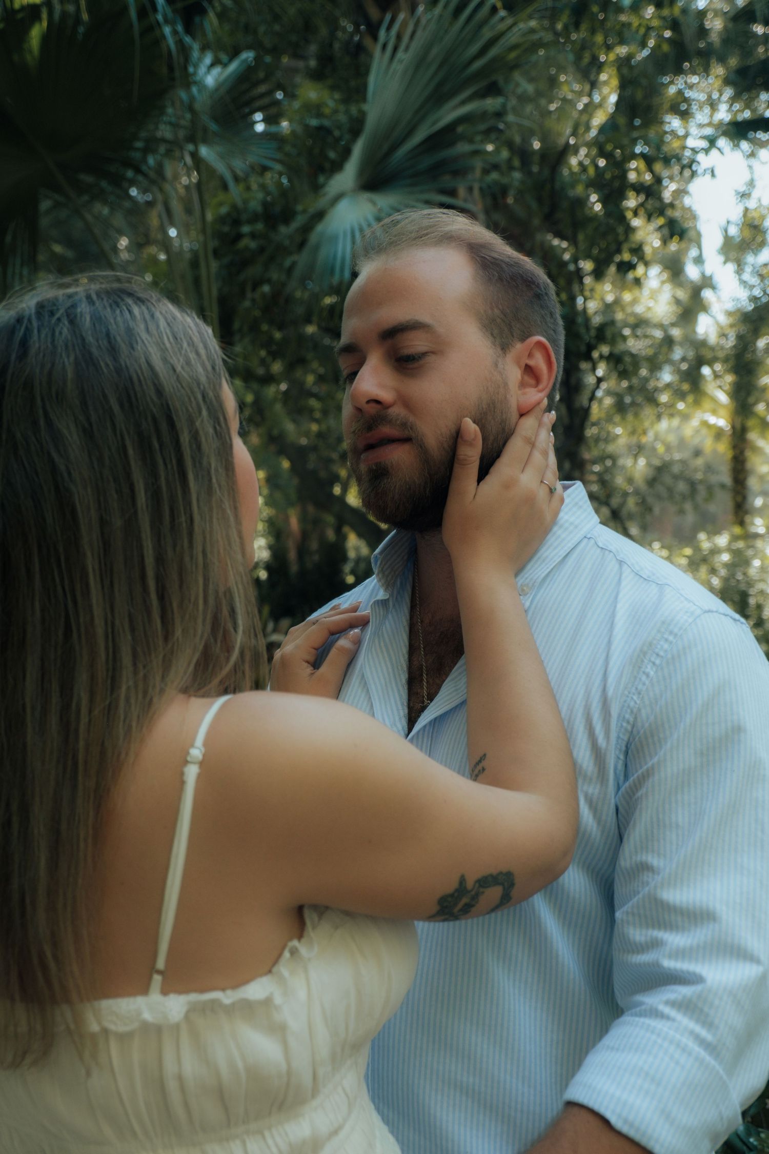Woman tenderly touches bearded mans face in romantic outdoor portrait surrounded by lush greenery.