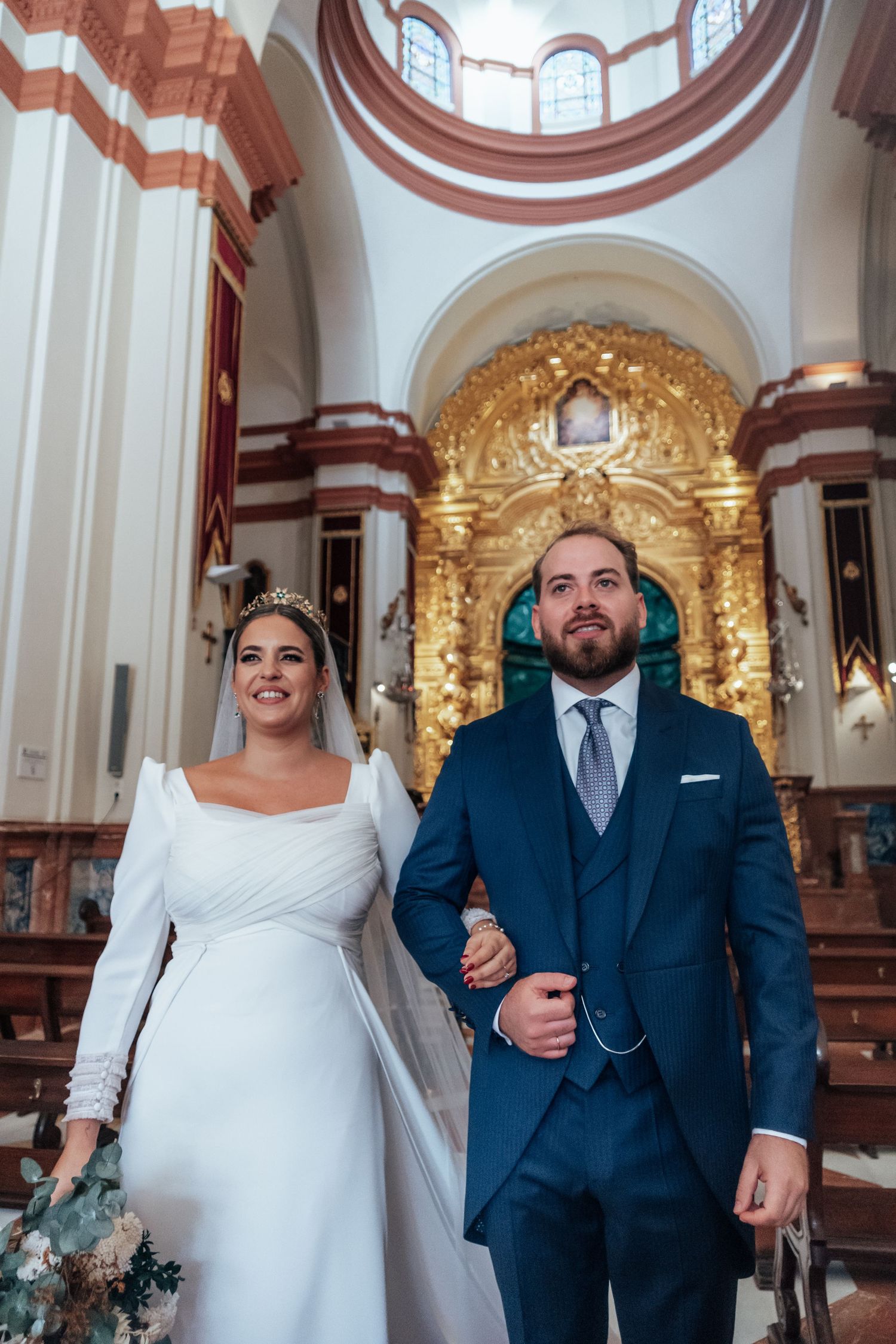 Newlyweds walk down church aisle, bride in white dress holding bouquet, groom in navy suit.