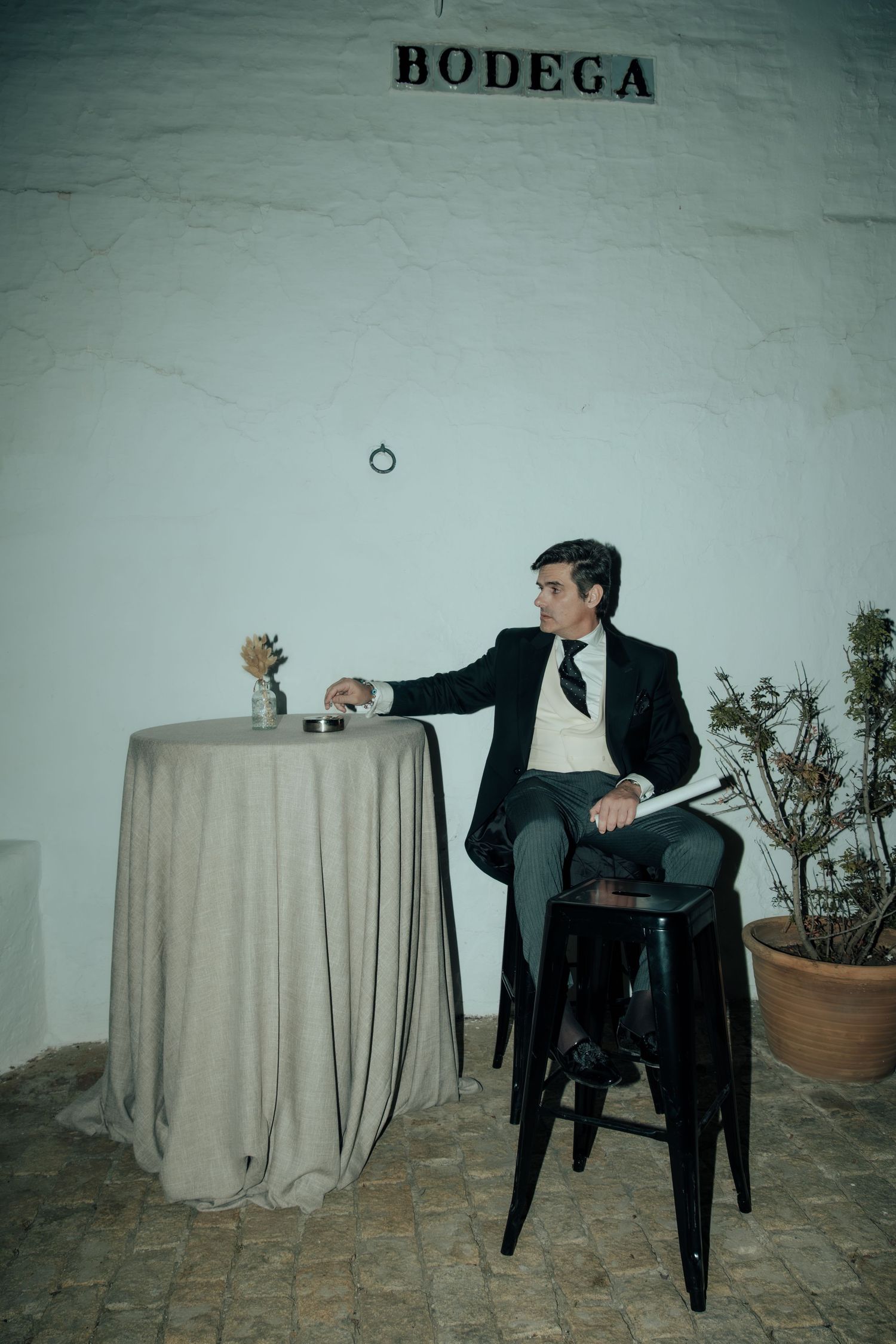 Elegant man in black suit sitting on stool beside draped table outside Bodega venue.