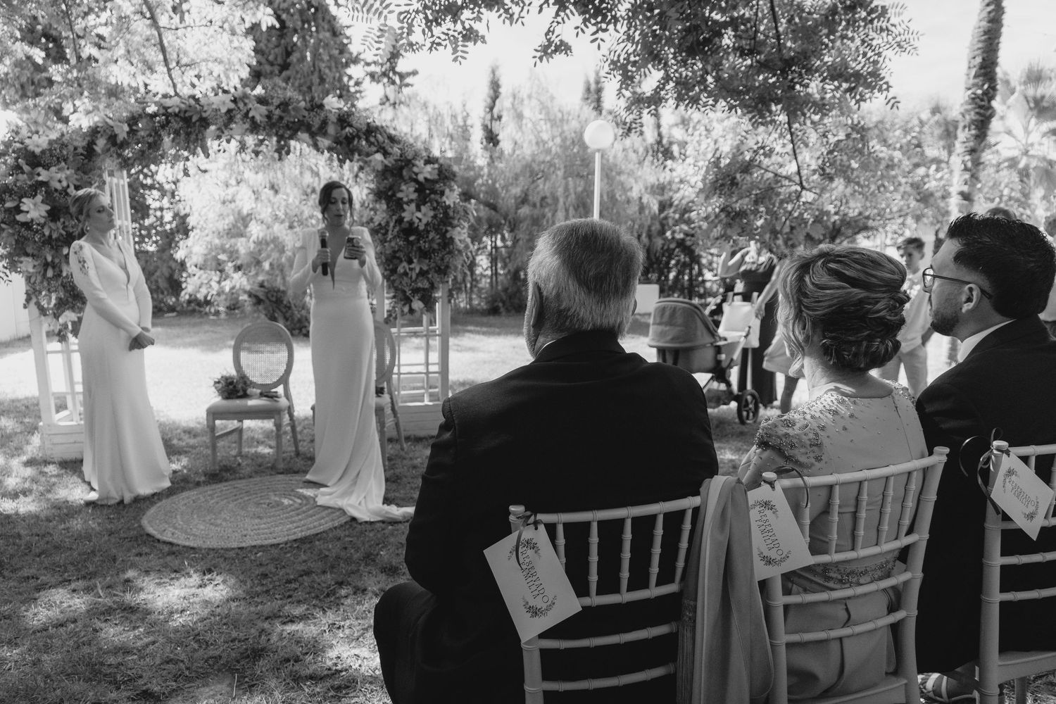 Black and white photo of two brides standing at floral arch during outdoor garden wedding ceremony.