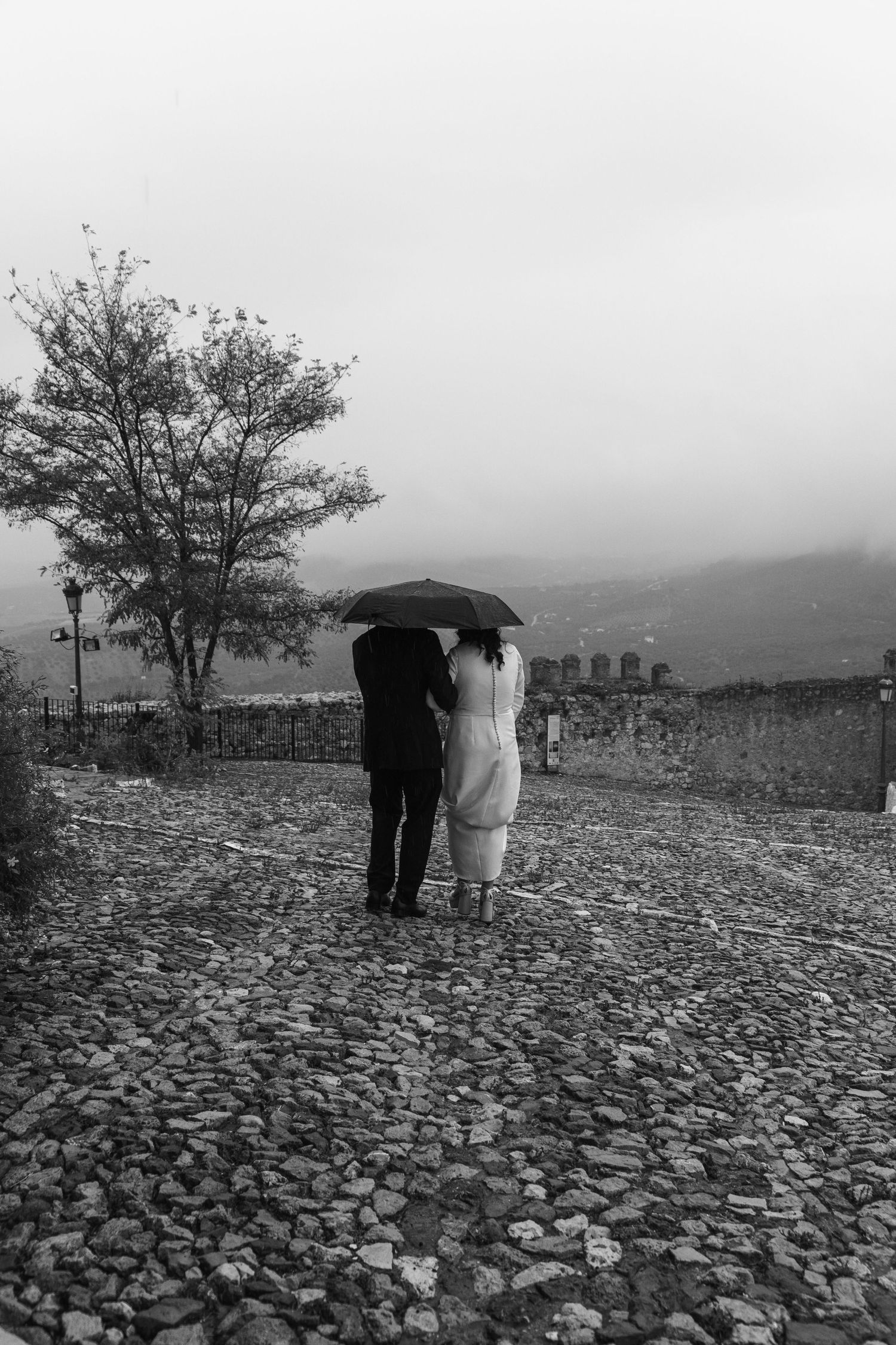Black and white photo of a newlywed couple walking together under an umbrella on a rainy cobblestone path.