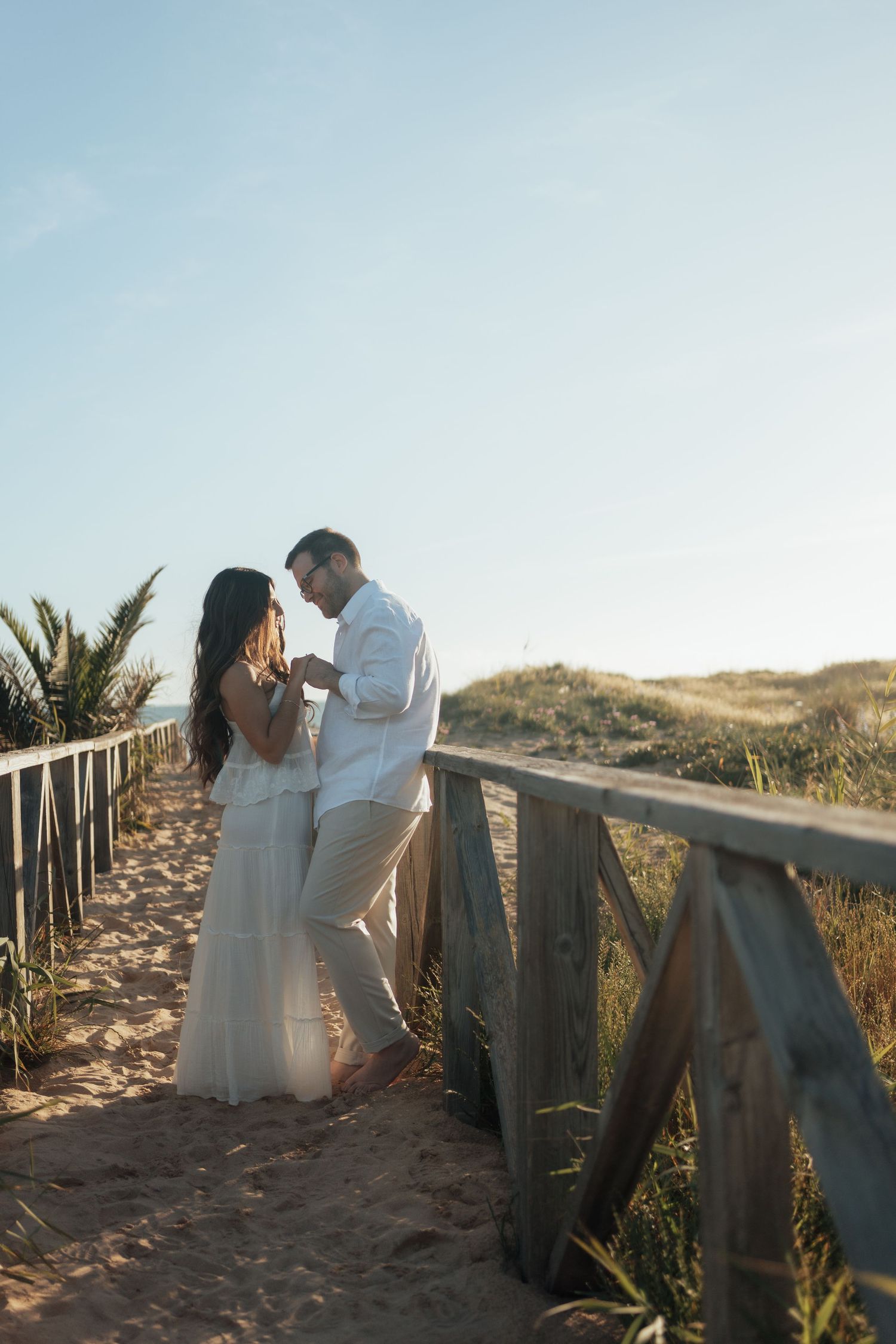 Romantic couple holding hands on a beach boardwalk surrounded by sand dunes at sunset.